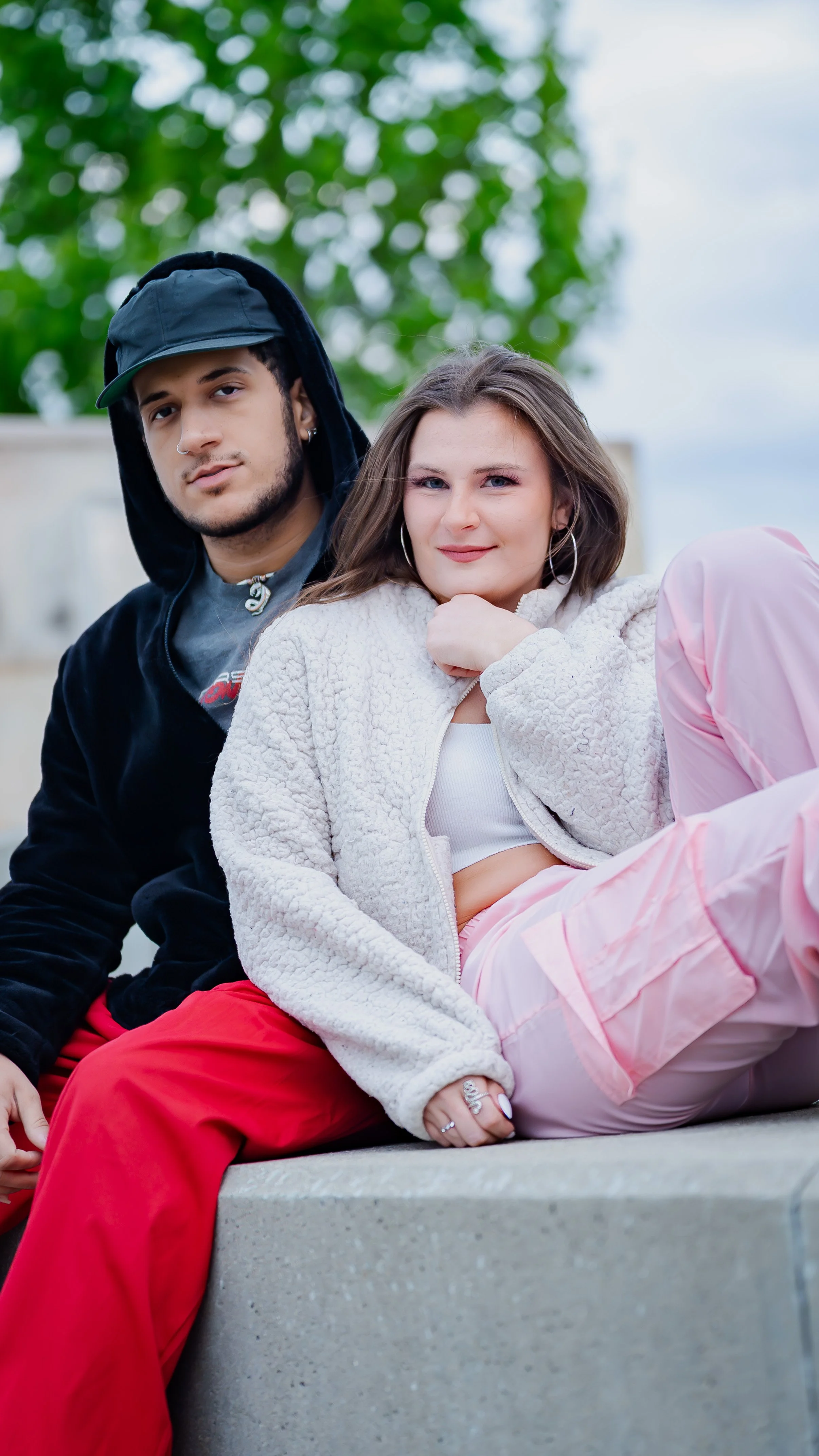 A young man and woman sitting on a concrete surface outdoors. The man has a nose piercing, is wearing a black hoodie with the hood up and a black cap, and has a neutral expression. The woman has long brown hair, is dressed in a light-colored sherpa j