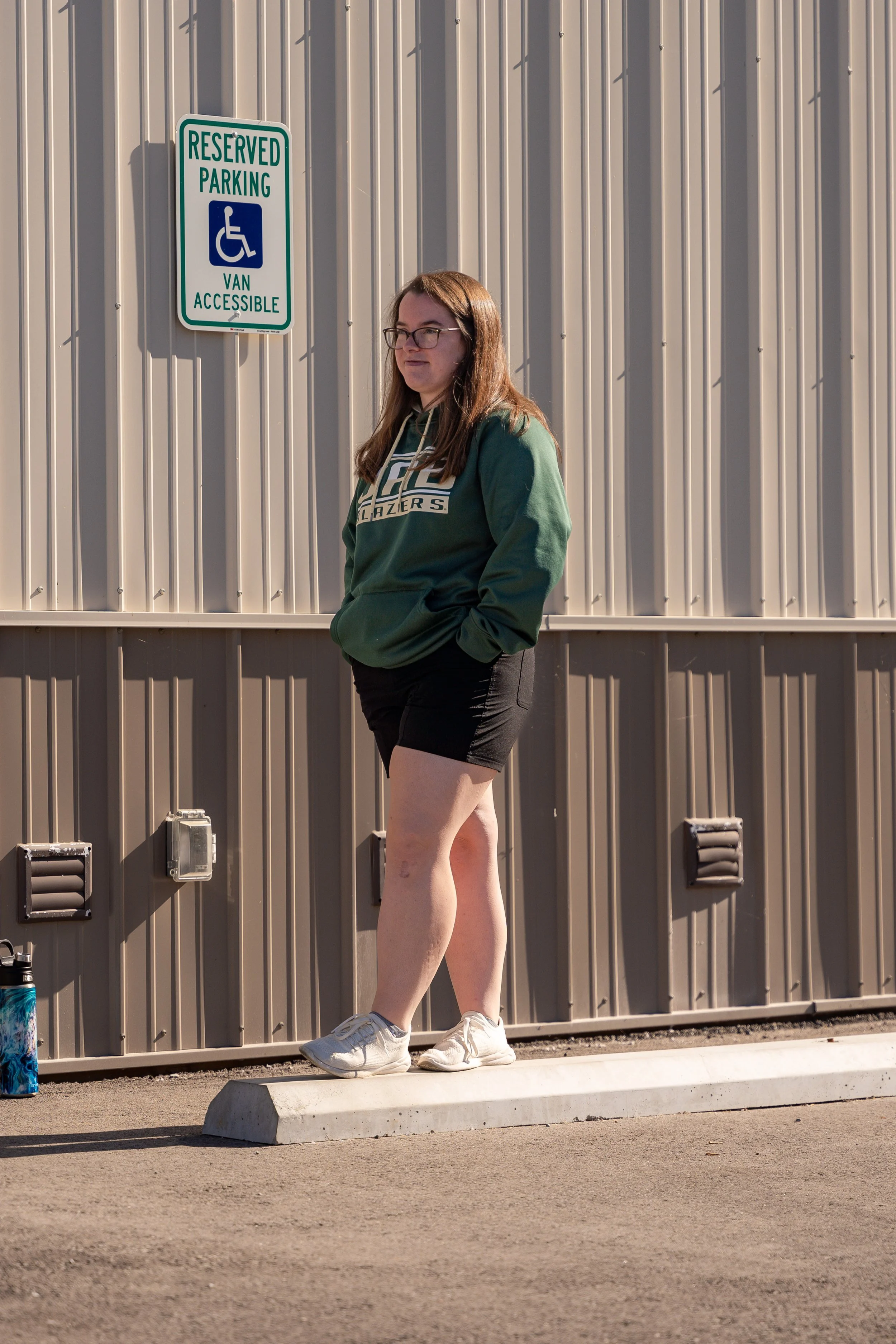 A young woman with long brown hair, glasses, wearing a green hoodie and black shorts, standing outdoors next to a metal building with a sign that reads Reserved Parking Van Accessible, with a person in a wheelchair symbol