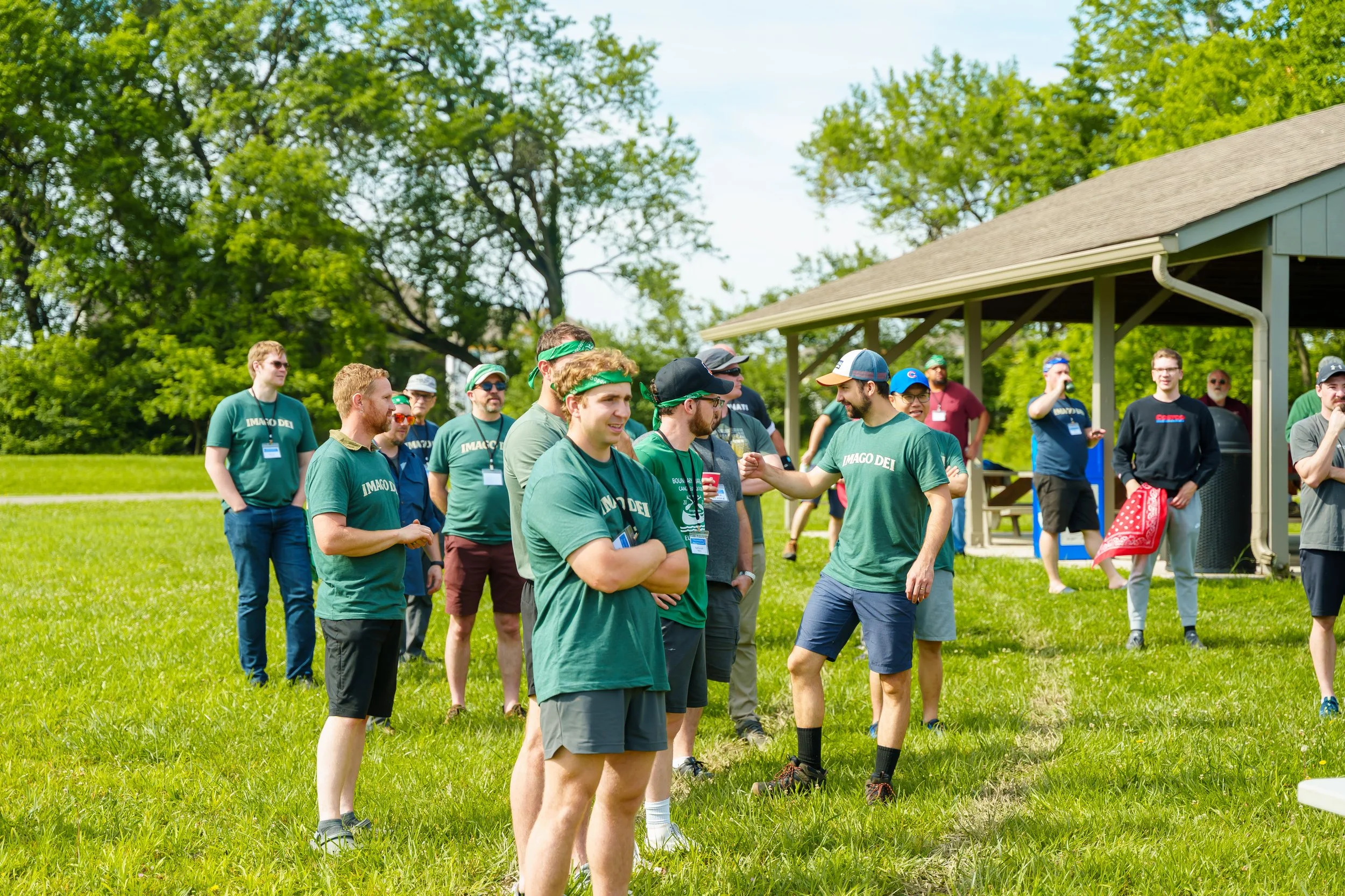 Group of young men and women outdoors on a sunny day, some wearing green shirts and bandanas, standing on a grassy field near a pavilion, engaging in conversation and socializing.