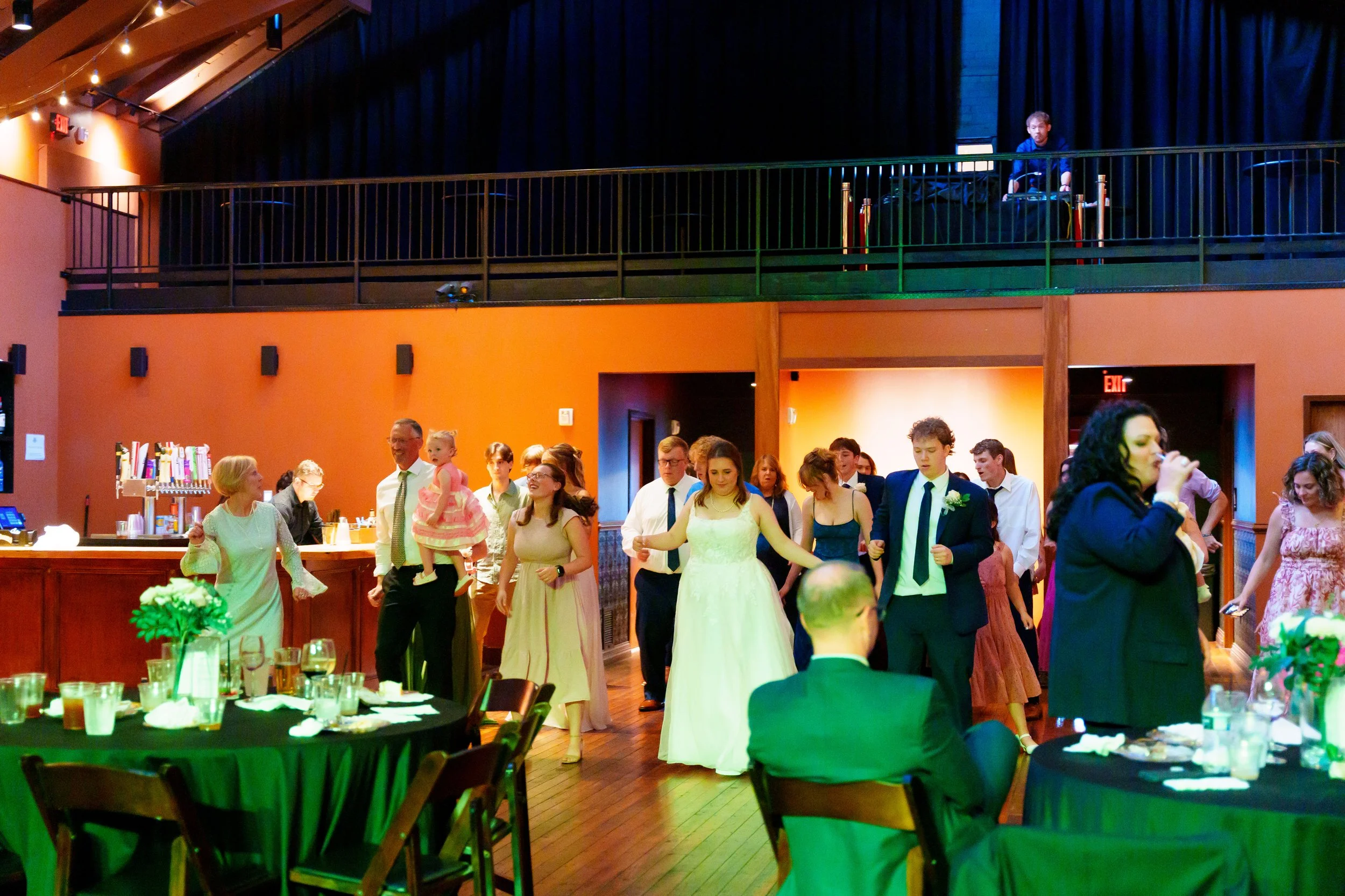 People dancing and socializing at a wedding reception inside a large hall with wooden floors and orange walls, tables with green tablecloths, and a bar area in the foreground.