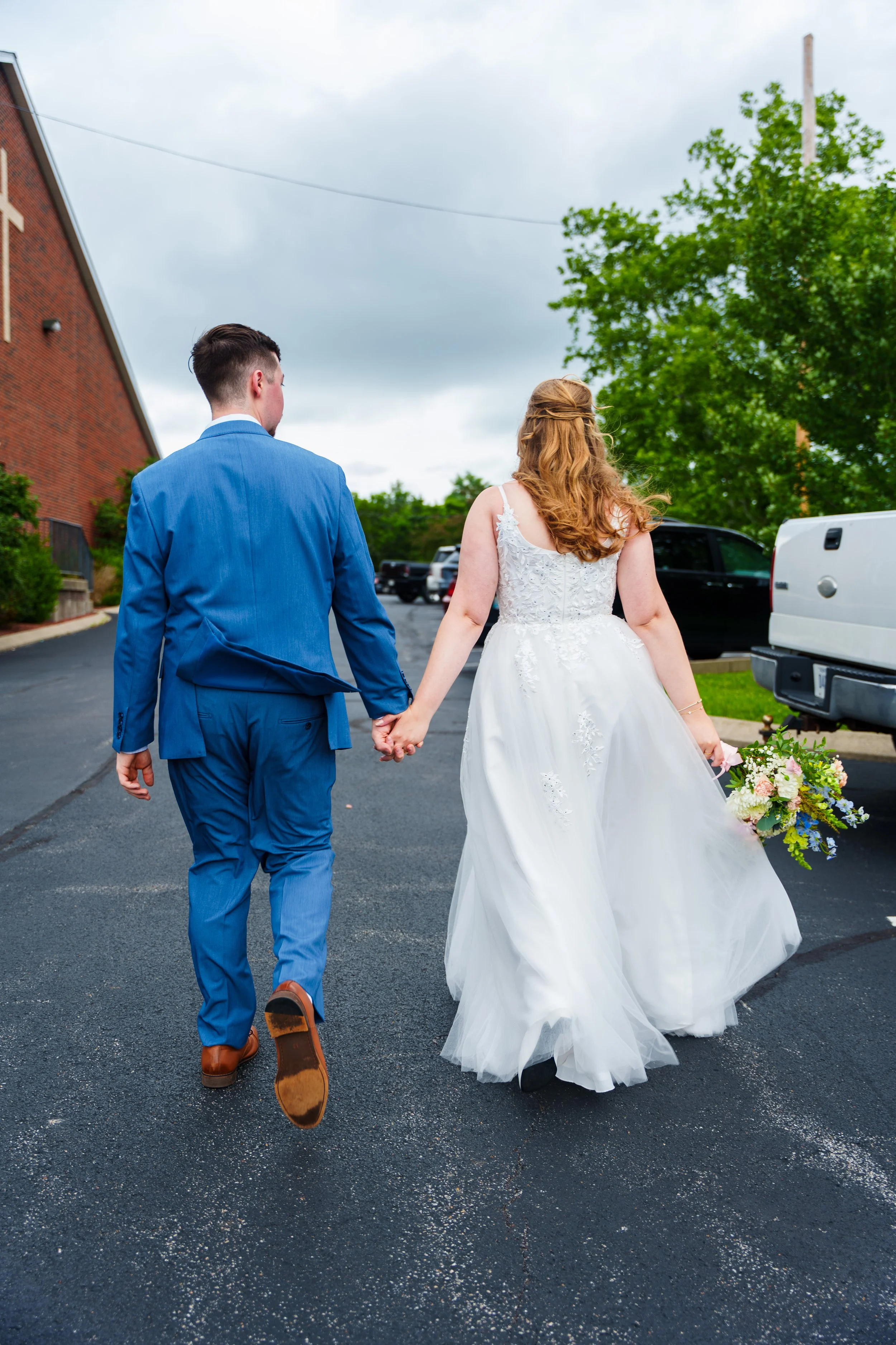 A bride and groom walking hand in hand outdoors on a cloudy day, with the groom wearing a blue suit and the bride in a white wedding dress holding a colorful bouquet.