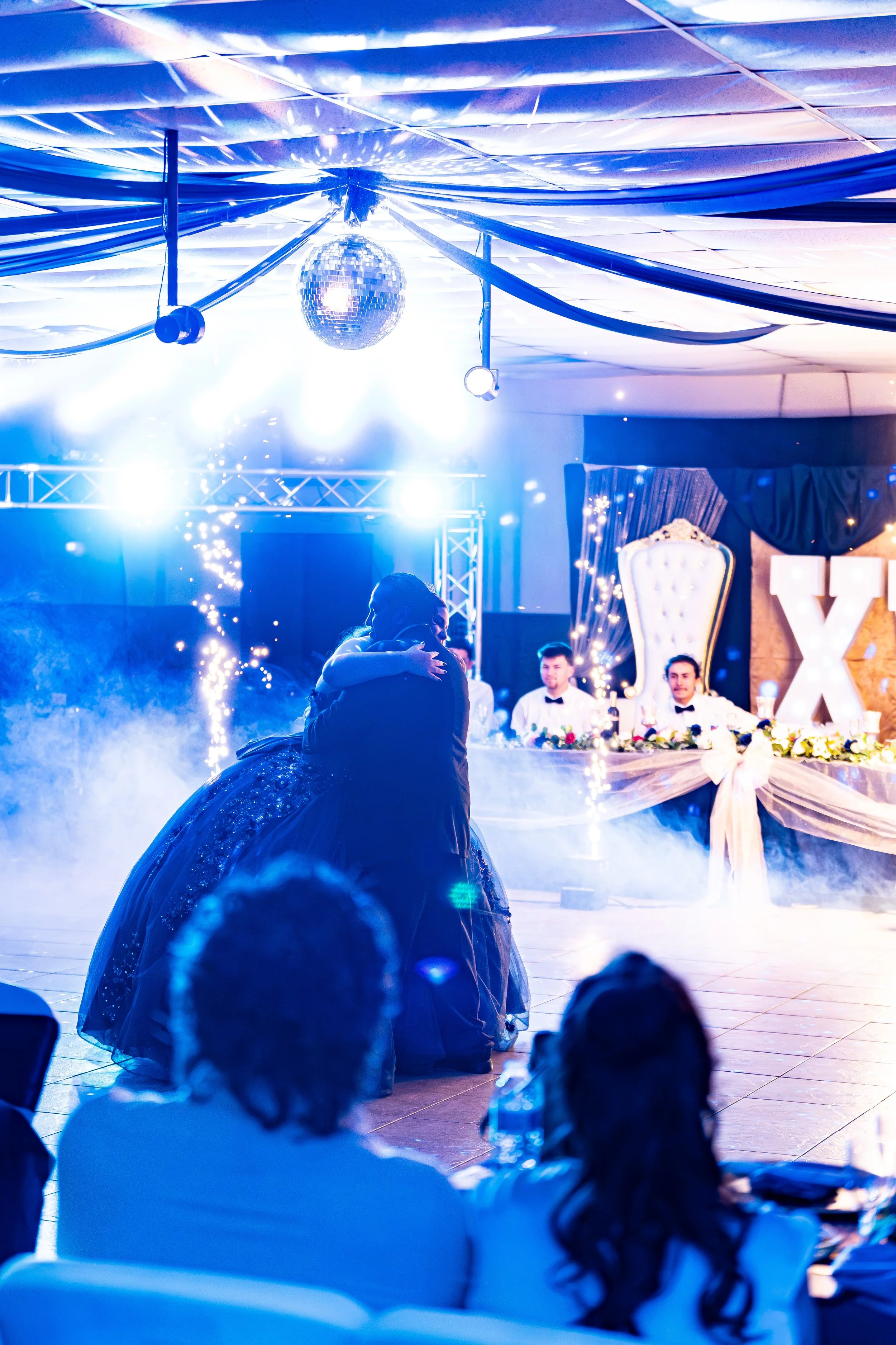 A couple in formal attire dancing at a party with a large lit-up sign, drapery, a disco ball, and sparklers in the background.