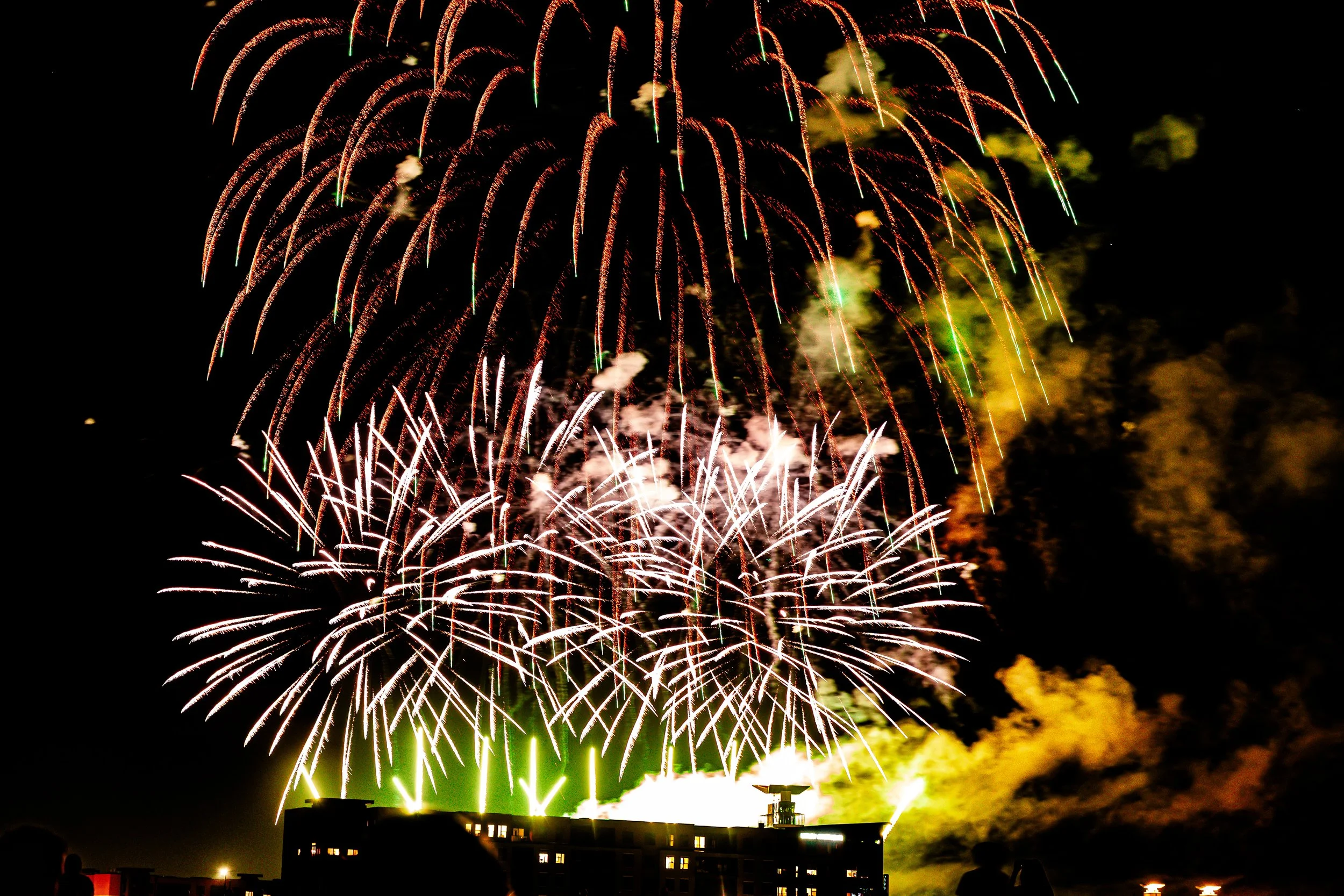 Fireworks display lighting up the night sky over a cityscape, with silhouettes of people watching from below.