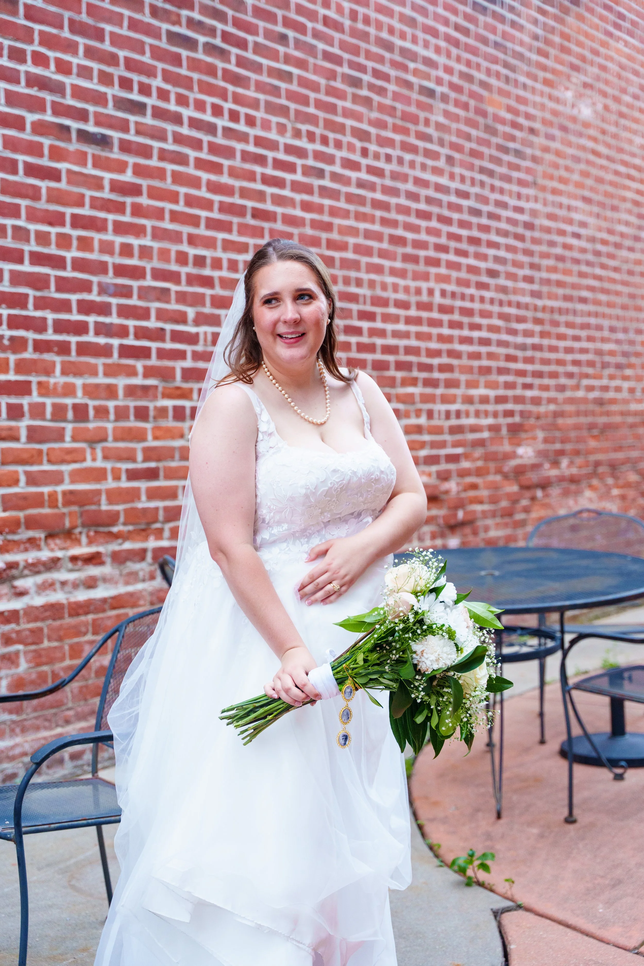 A bride in a white wedding dress holding a bouquet of white flowers and greenery, standing outdoors against a red brick wall.