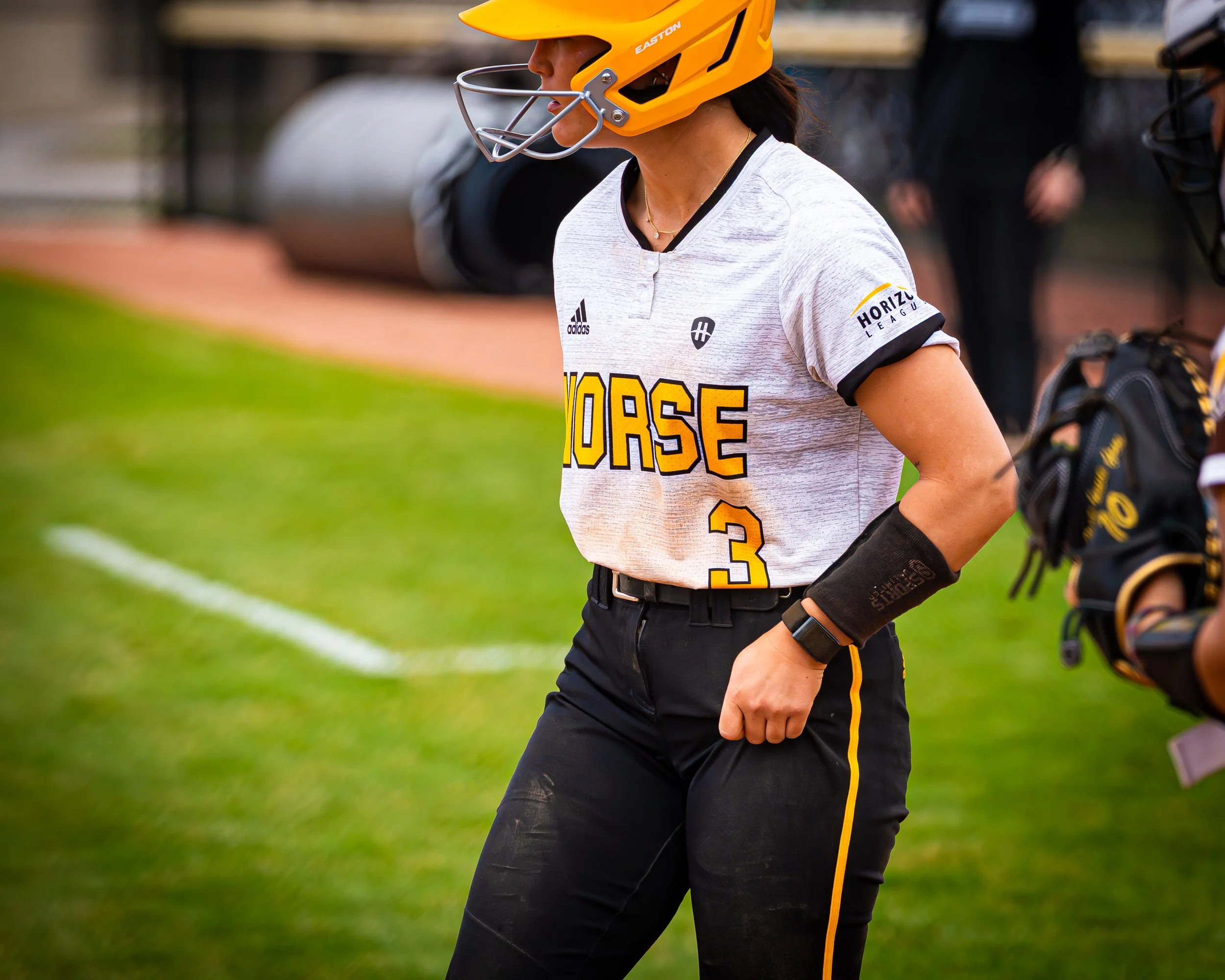 A female baseball player wearing a yellow helmet and white baseball jersey with black and yellow accents, standing on a grassy field. She is holding a black baseball glove and has her hands on her hips, with a row of baseball bats in the background.
