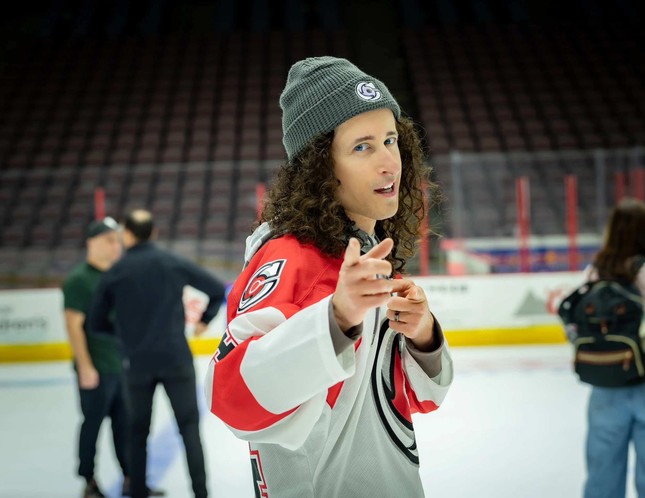 Person with curly hair wearing a gray beanie and a hockey jersey on an ice hockey rink.