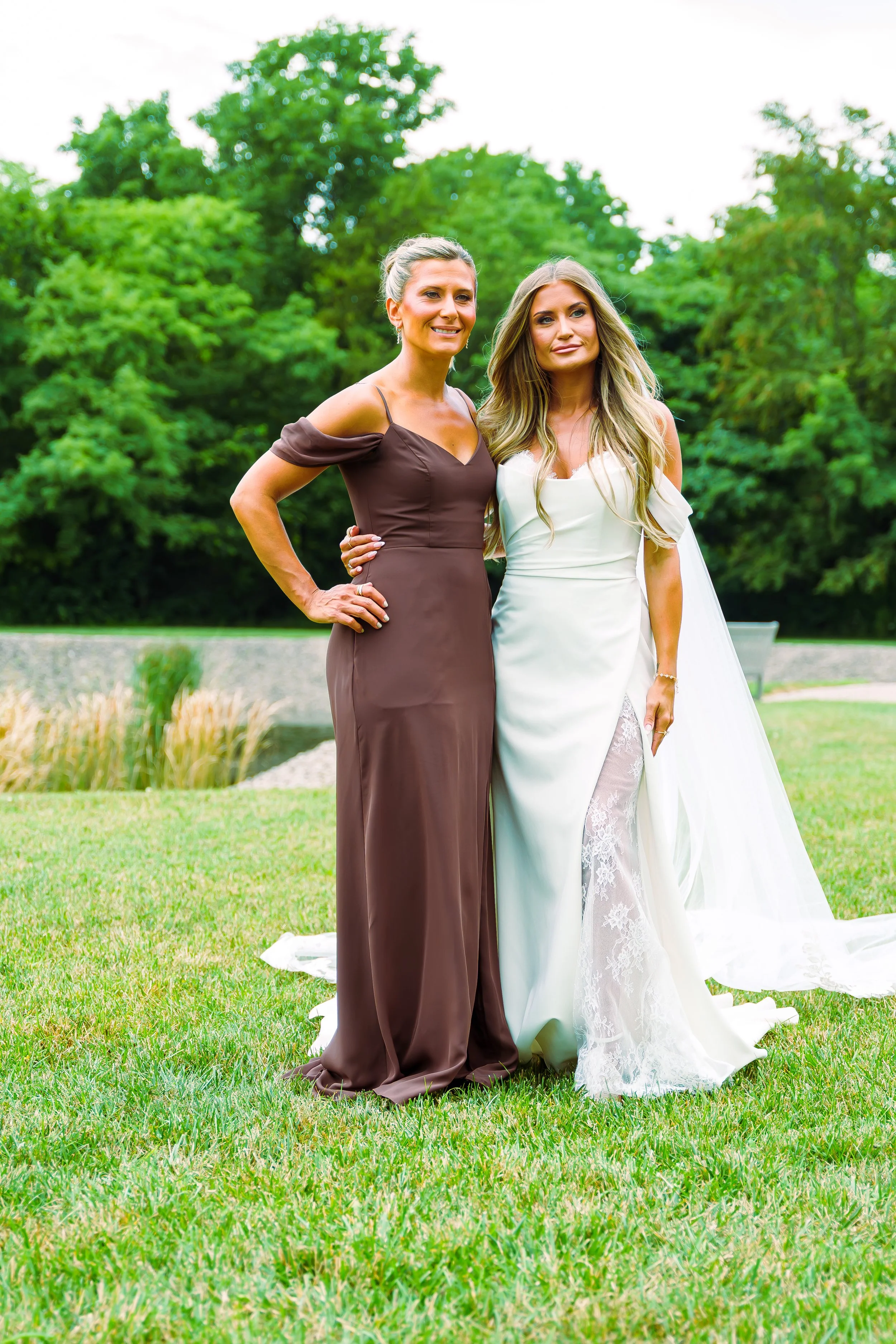 Two women stand outdoors on a grassy area with trees in the background, one in a brown dress and the other in a white wedding gown, posing for a photo.