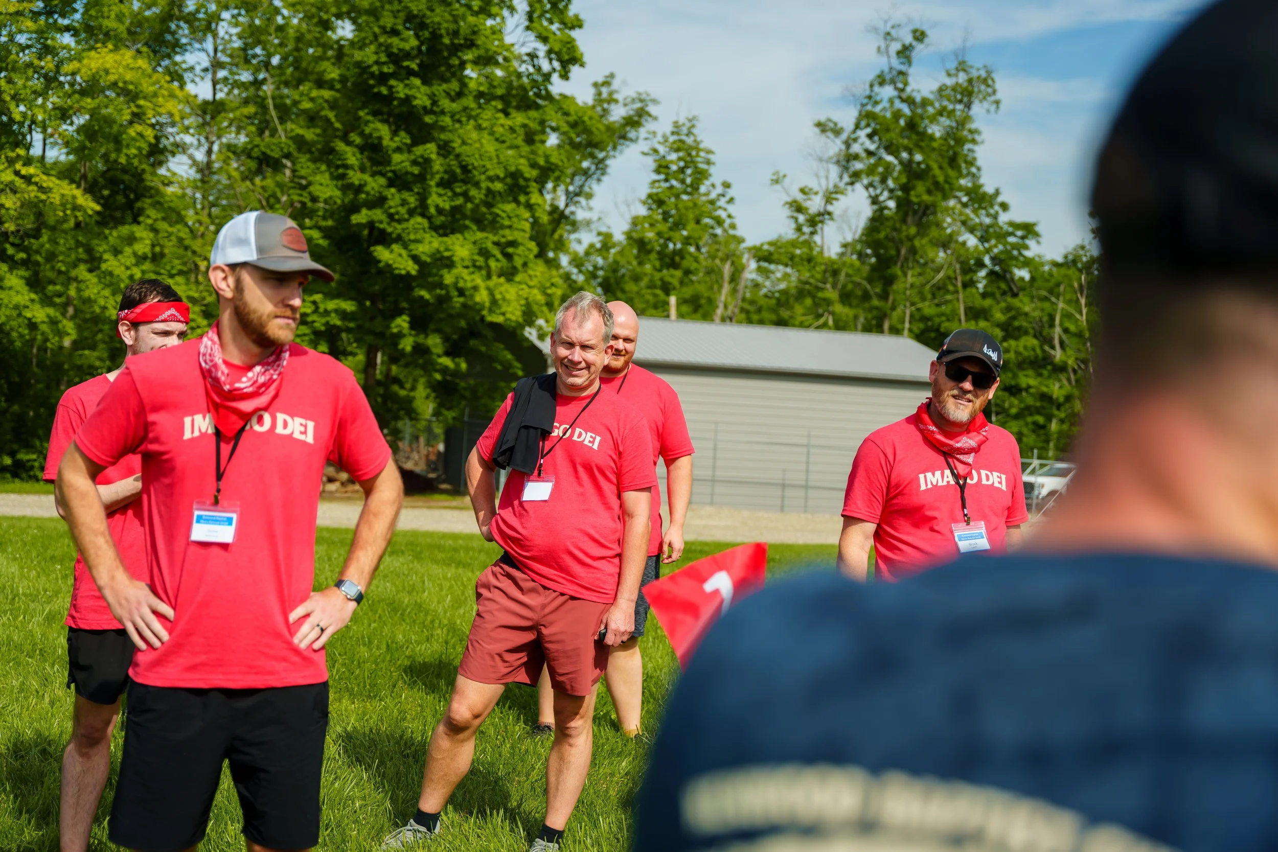 Group of people wearing red shirts and red bandanas gathered outdoors on a grassy area with trees in the background, participating in an event or activity.