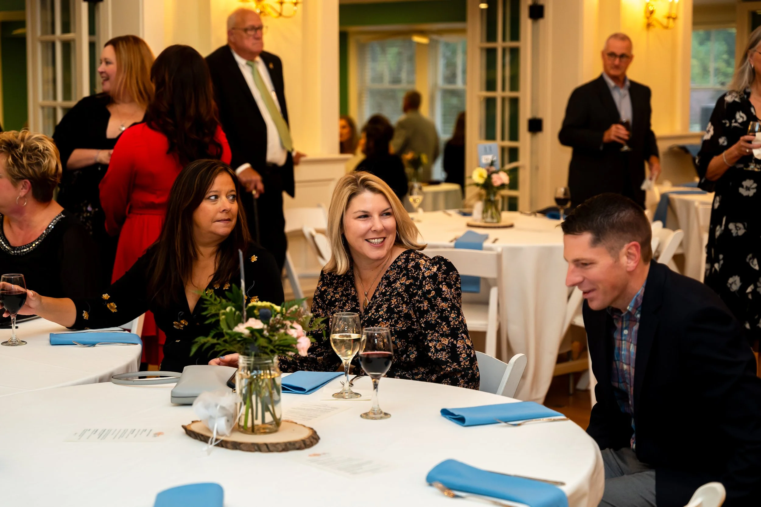People gathered at a round dining table with blue napkins, wine glasses, and floral centerpieces in a warmly lit room, smiling and engaging in conversation.