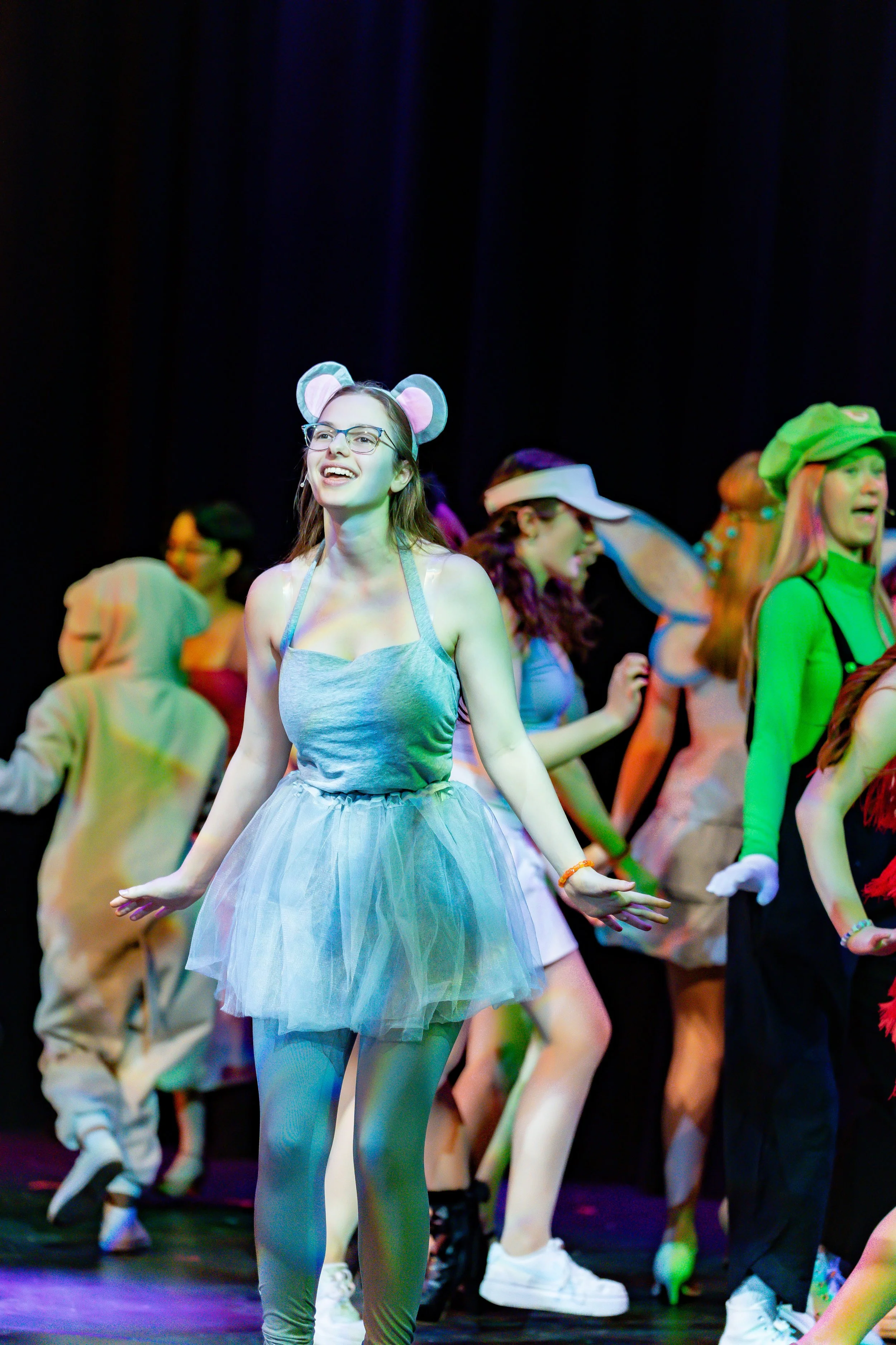 Young woman with glasses dancing on stage surrounded by other colorful performers, wearing mouse ears headband and a tutu.