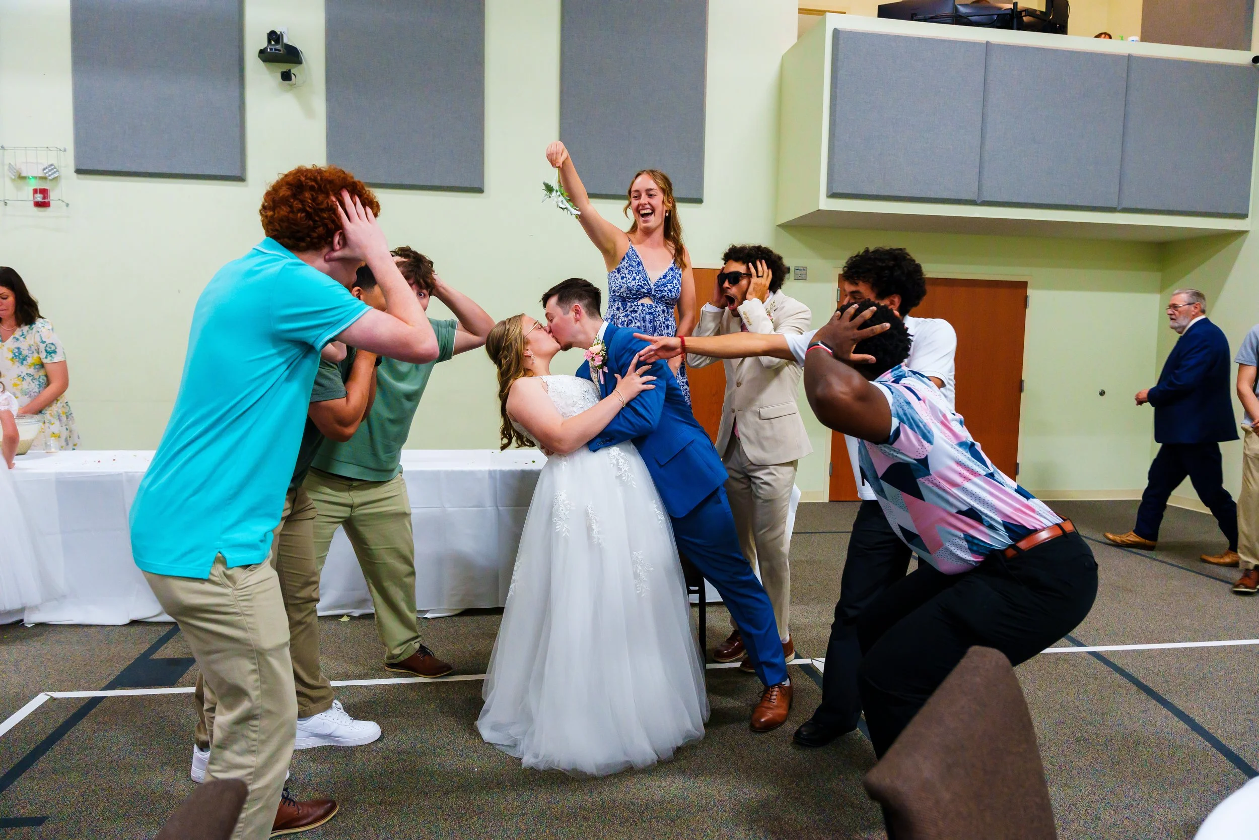 A group of young adults celebrating at a wedding reception, with a bride and groom kissing in the center, surrounded by friends who are cheering, laughing, and showing excitement.