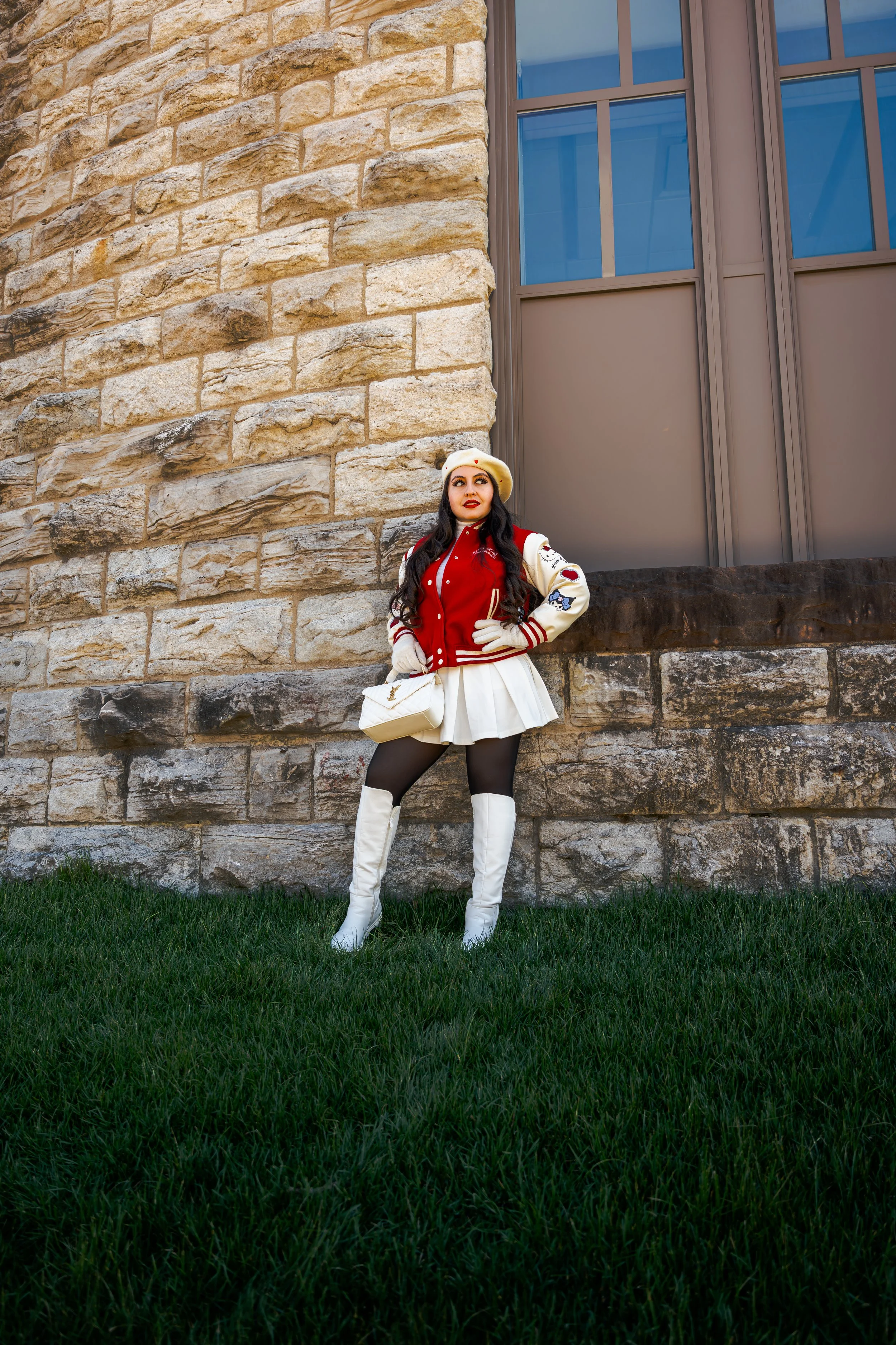 A woman with long dark hair, wearing a beige beret, red and white varsity jacket, white skirt, black tights, and white knee-high boots, standing outdoors against a stone wall with a large window, holding a white purse.