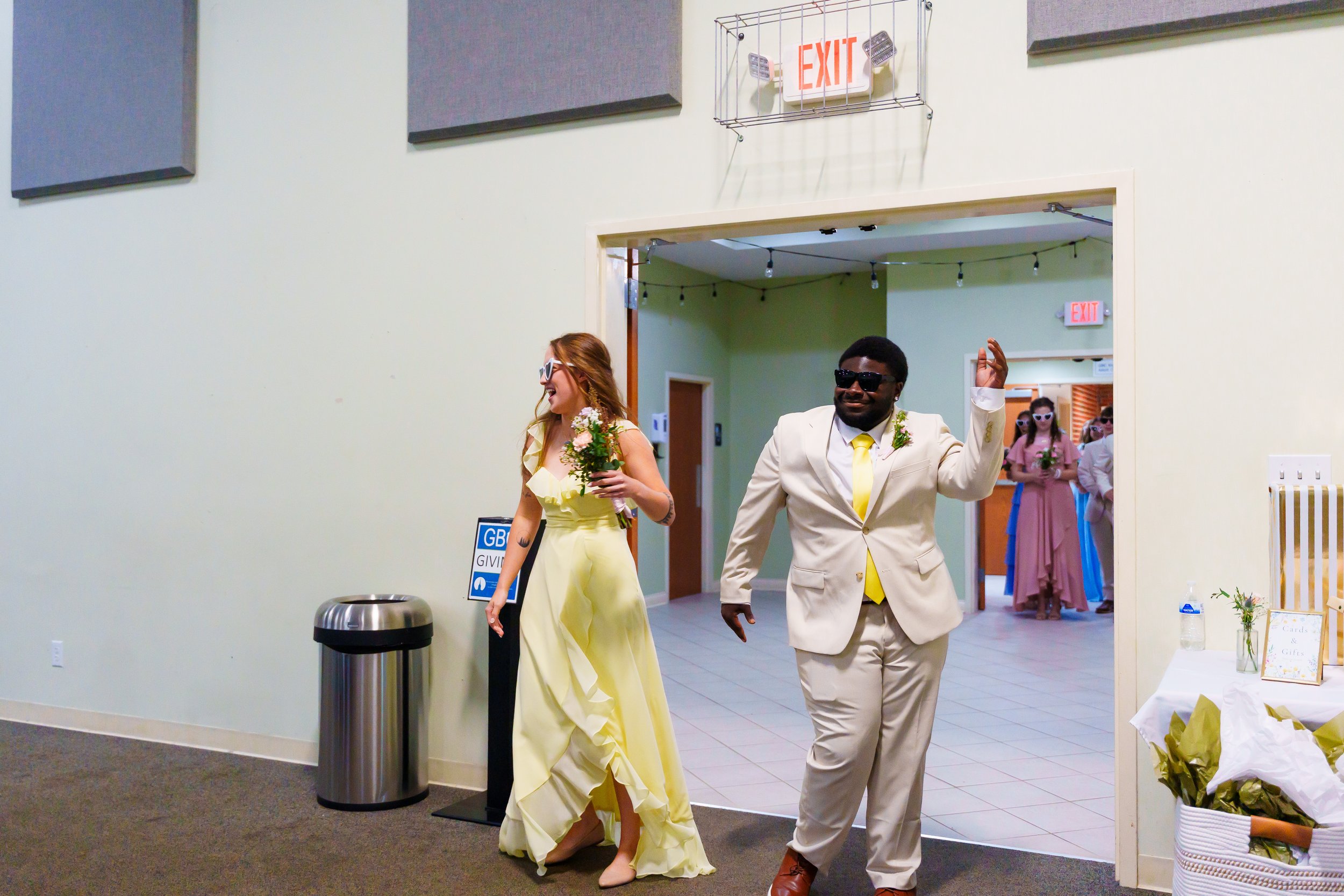 A woman in a yellow dress holding a bouquet and wearing sunglasses, and a man in a beige suit with a yellow tie and sunglasses, are walking out of a room with happy expressions. Other women in pink dresses with sunglasses are in the background.