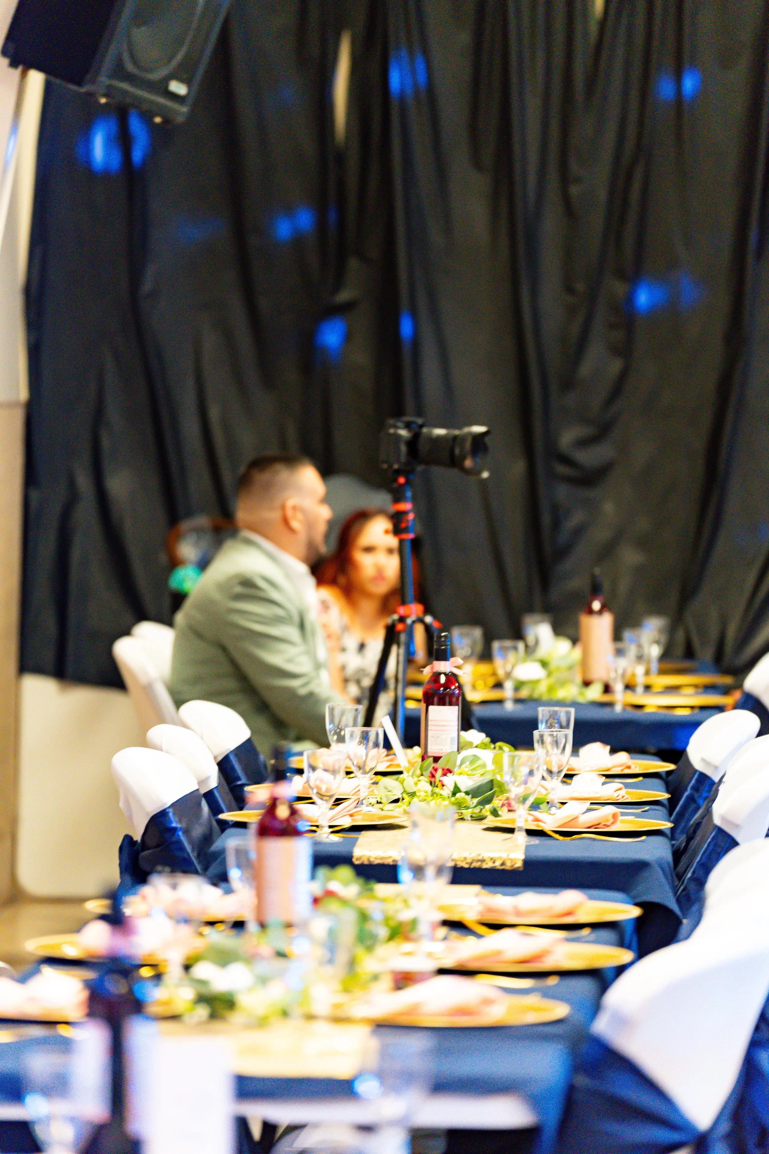 Table set for a celebration with yellow plates, floral centerpieces, wine bottles, glasses, and utensils. Guests seated, with a black curtain backdrop and blue lighting.
