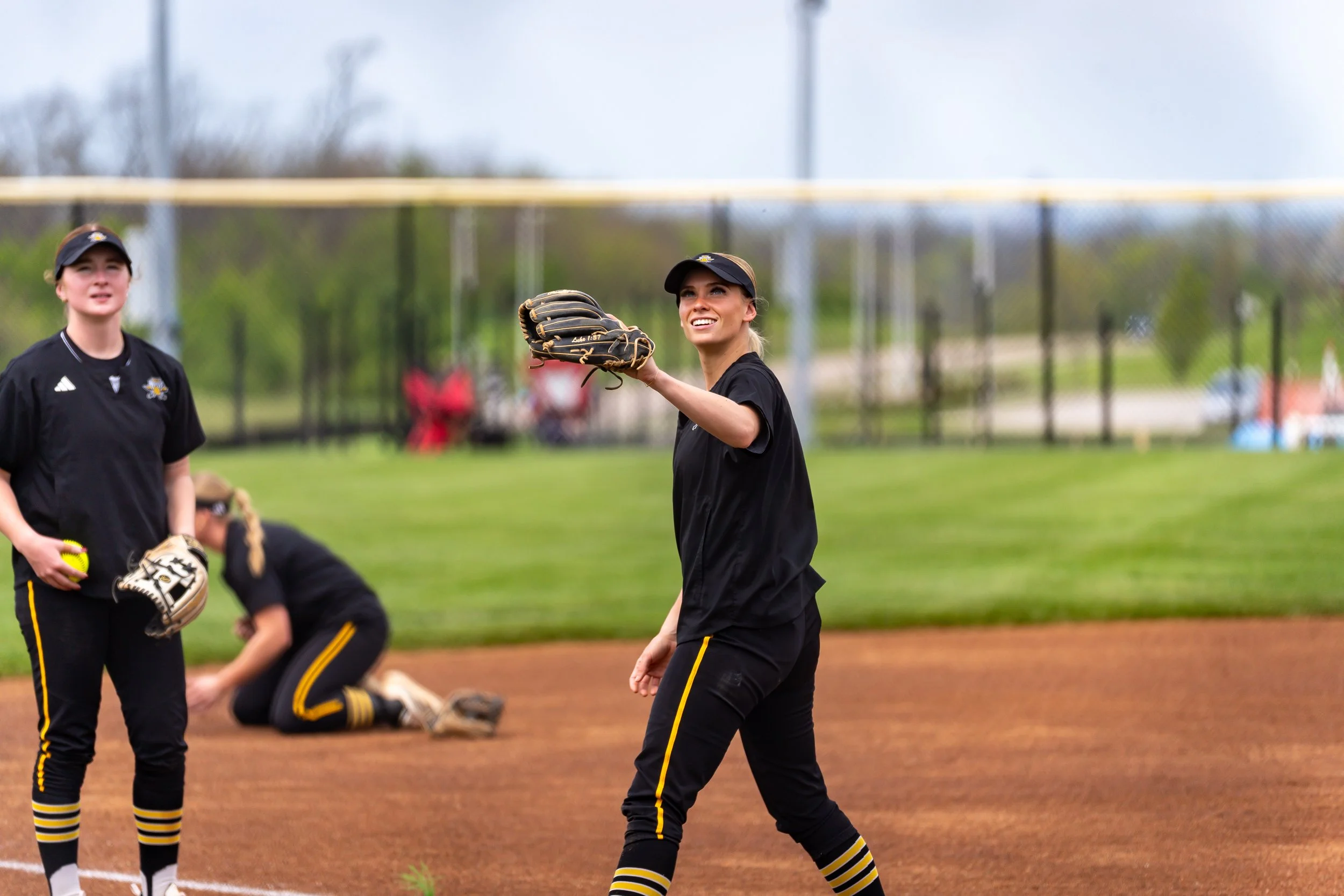 Softball players on the field, with one smiling and holding a glove, others preparing for play on a grassy and dirt softball field during daytime.