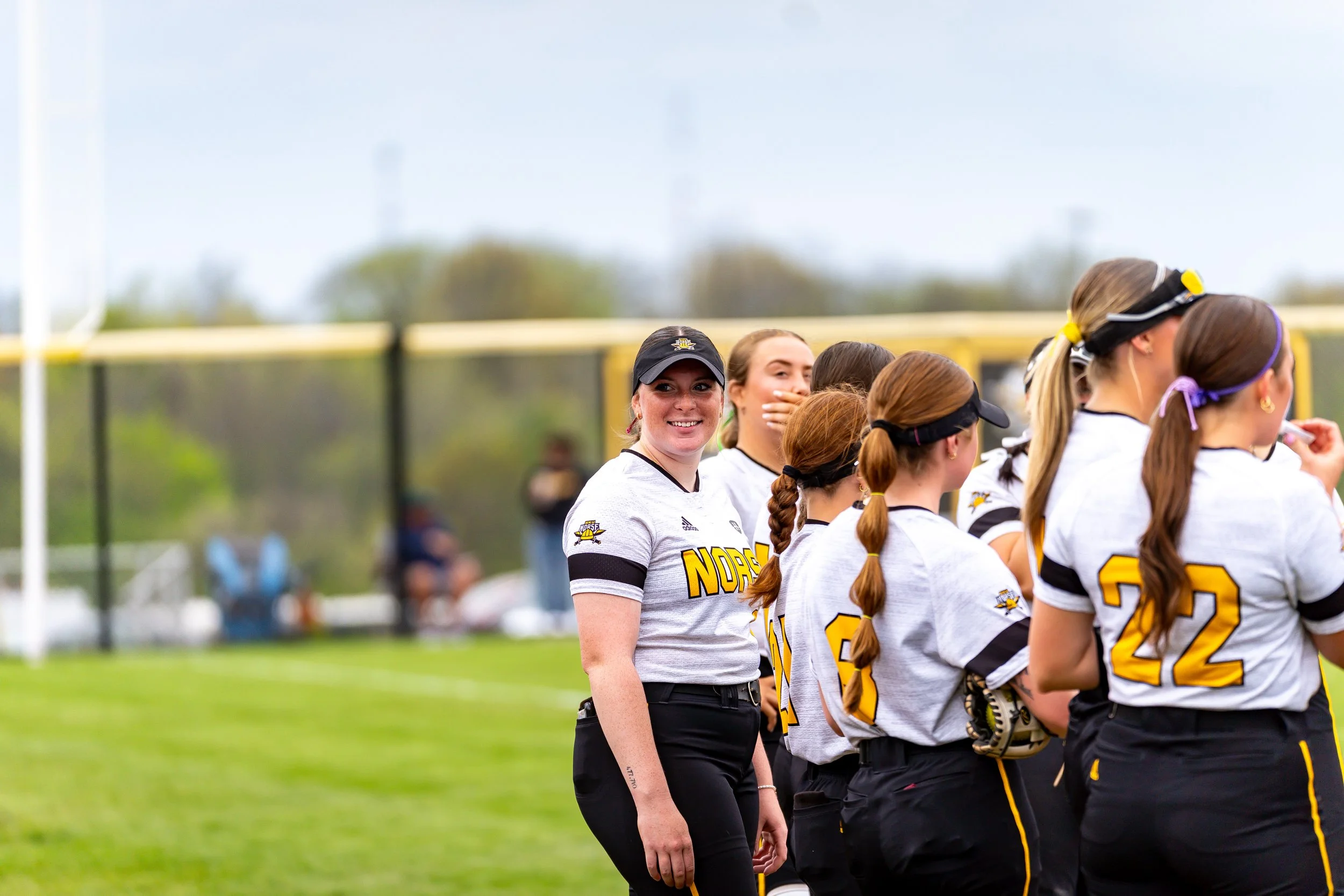 A group of female softball players in uniform standing on a field, with one player smiling at the camera in the foreground.