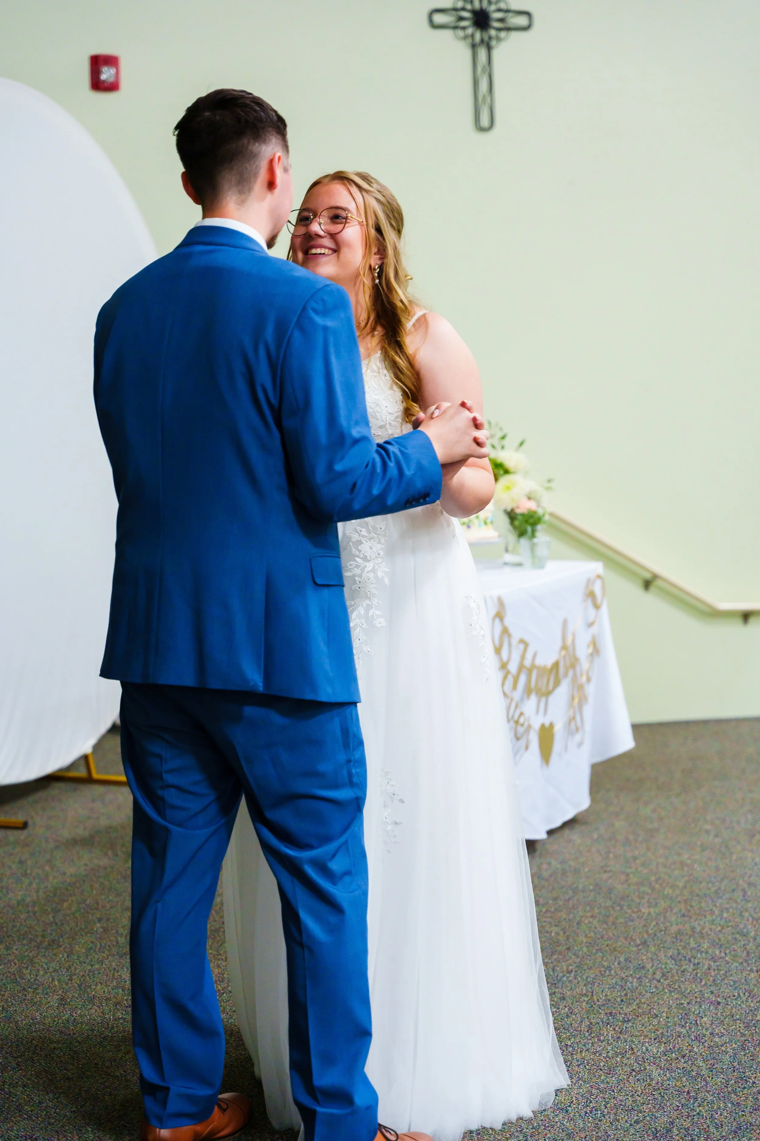 A couple dressed in wedding attire is holding hands and smiling at each other in a church, with a crucifix on the wall and a table with flowers and a 'Happy Wedding' banner in the background.
