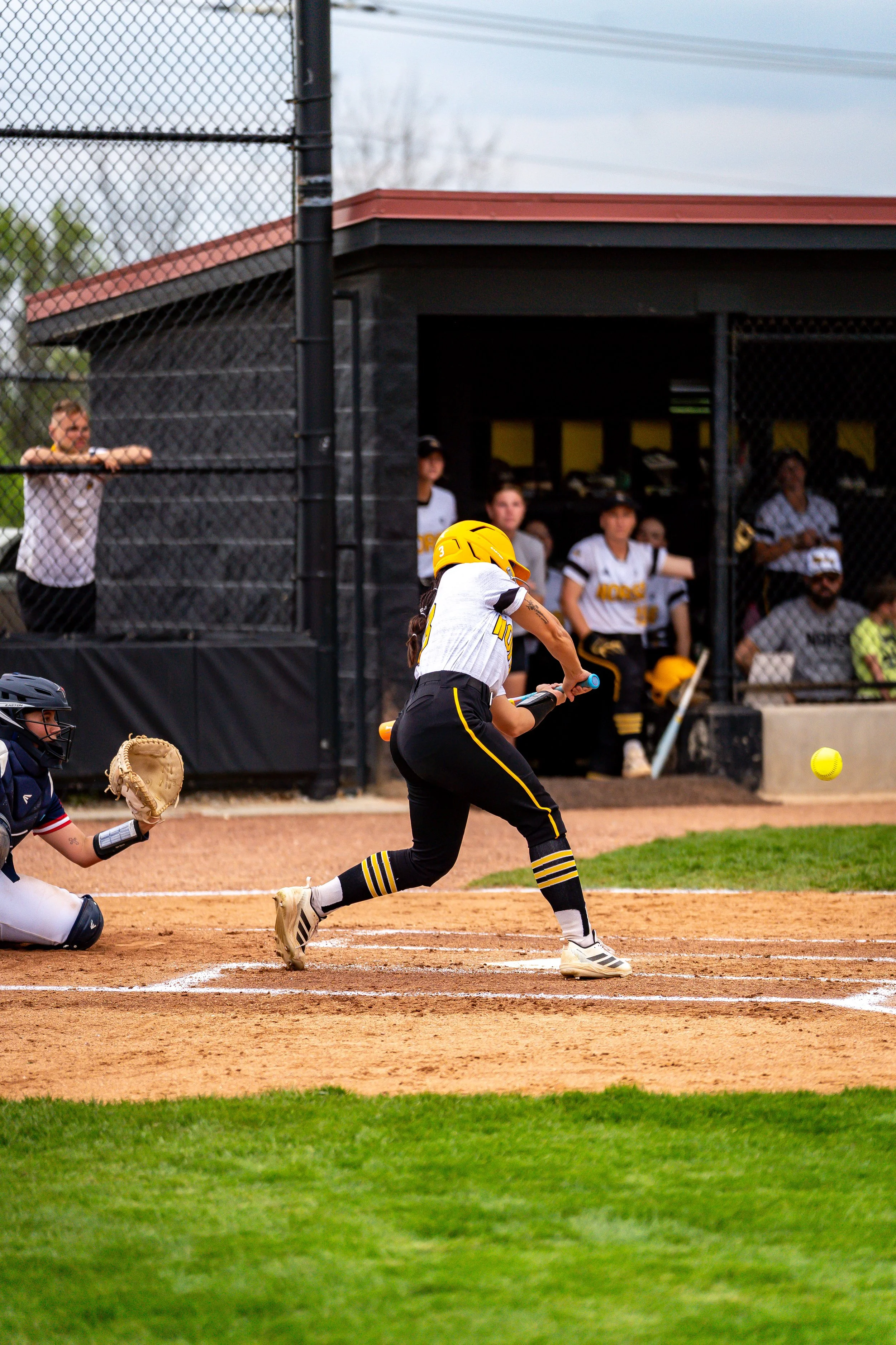 A female softball player in black and yellow uniform batting at home plate with a yellow softball approaching.