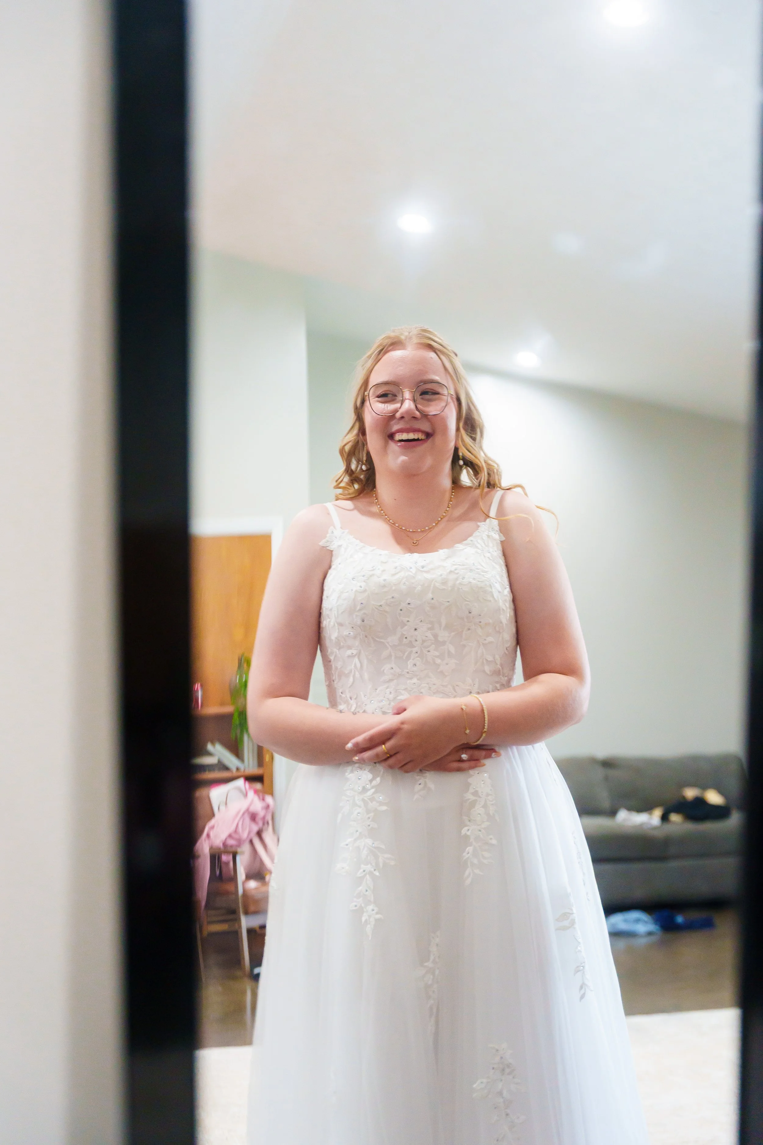 A woman with glasses and blonde hair, smiling, wearing a white lace dress, standing in front of a mirror, preparing for a special occasion.