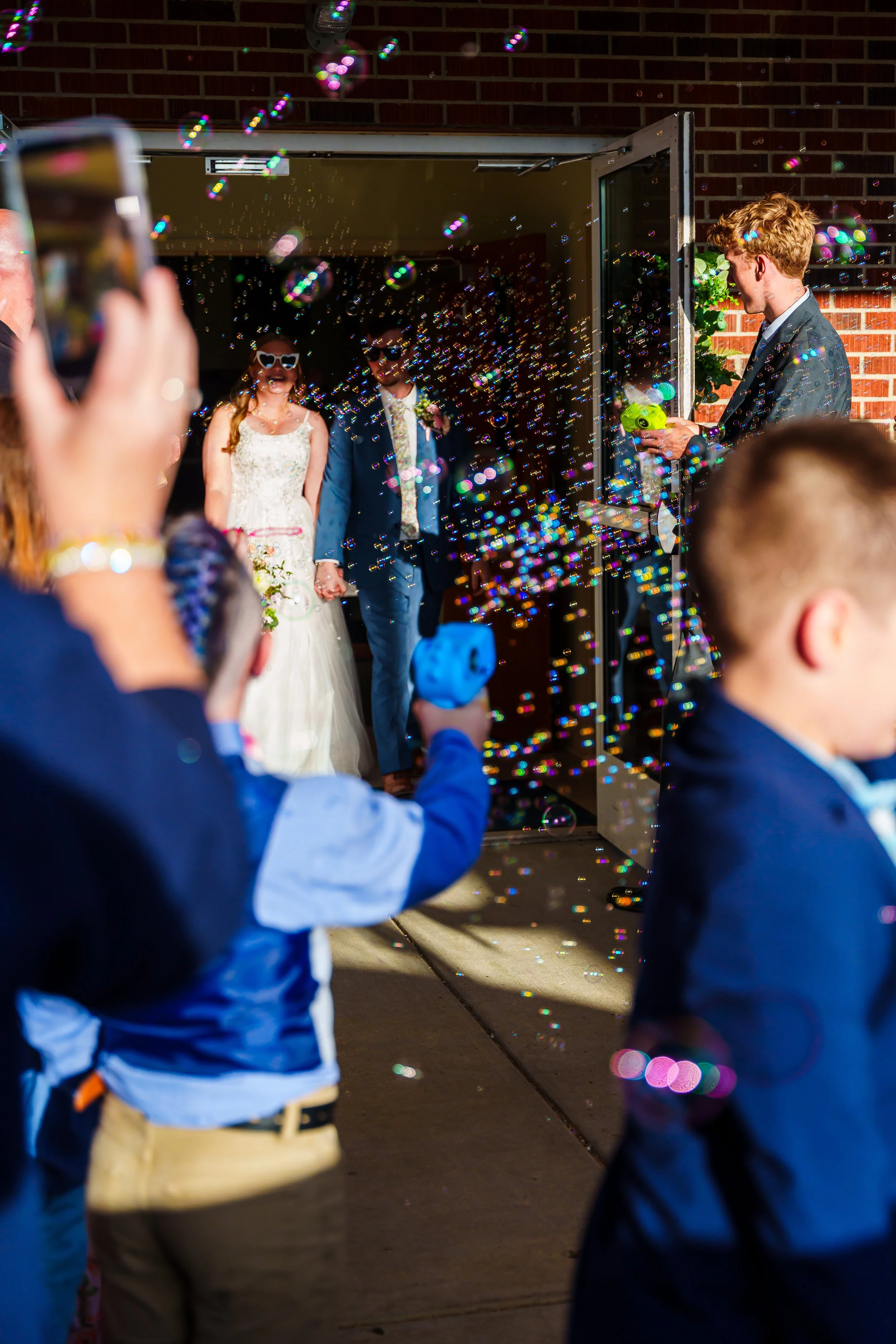 A wedding celebration with a bride and groom holding hands as they enter a venue, with guests in the foreground taking photos and children in suits playing with bubbles.