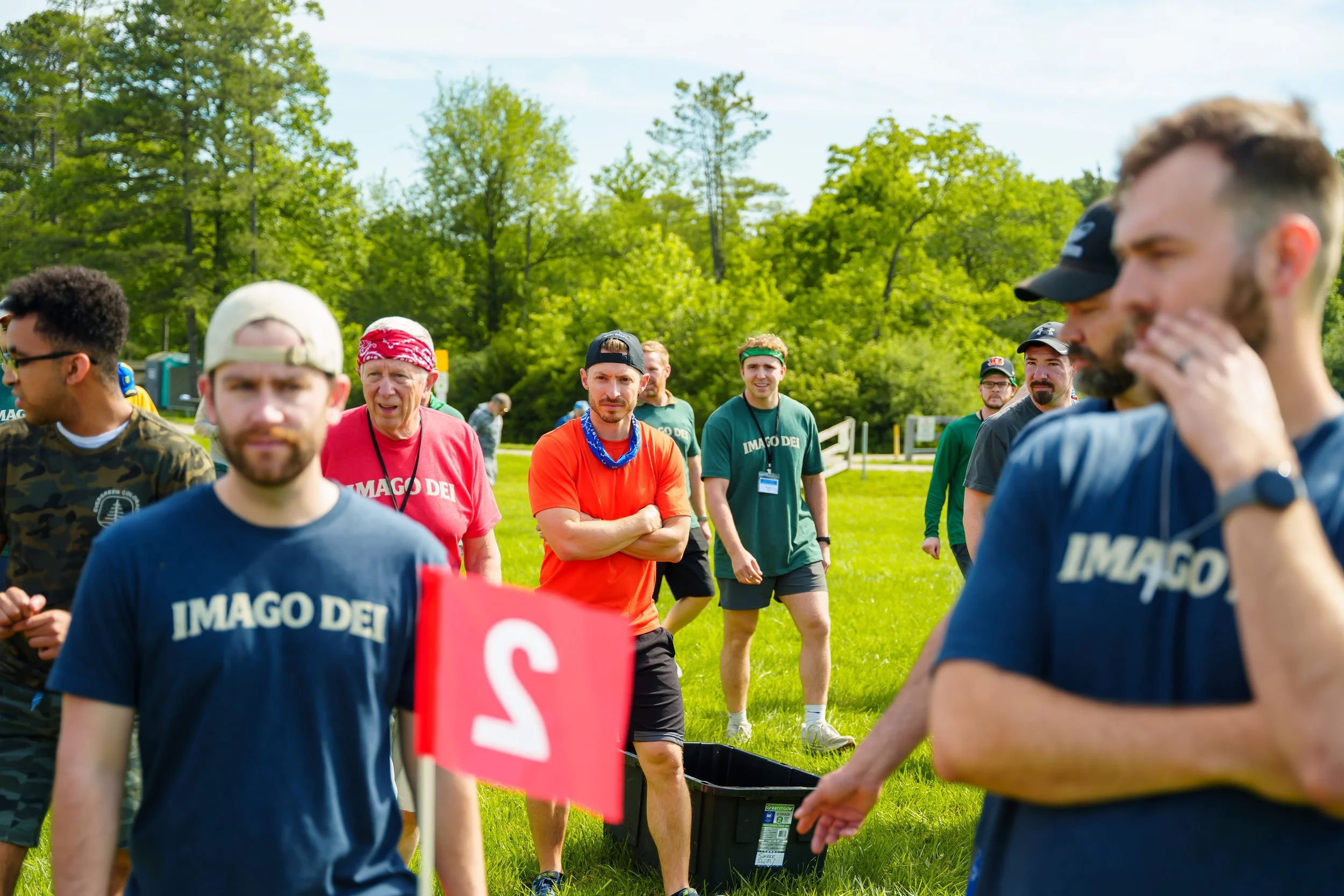 Group of people gathering outdoors on grassy field, some wearing green shirts with 'IMAGO DEI' printed on them, in a park with trees in the background. It appears to be a community event or team activity, with individuals standing around and interact