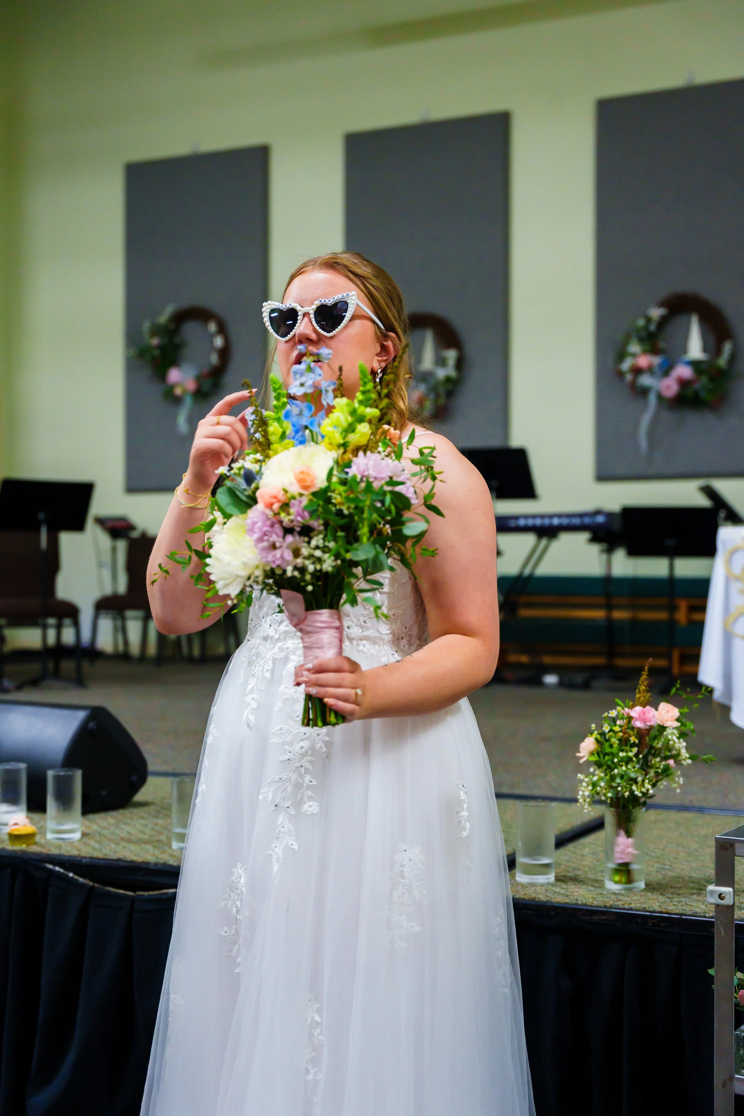 A bride in a white wedding dress holding a bouquet of flowers, wearing large heart-shaped sunglasses, standing indoors with floral wreaths hanging on the wall behind her.