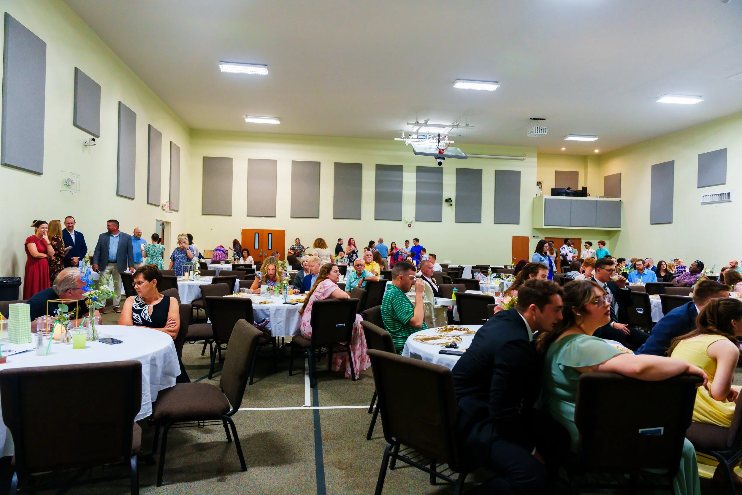 A large banquet hall filled with guests seated at round tables with white tablecloths, decorated with floral centerpieces, with people socializing and eating. The hall has high ceilings, numerous overhead lights, and a wall with gray soundproof panel