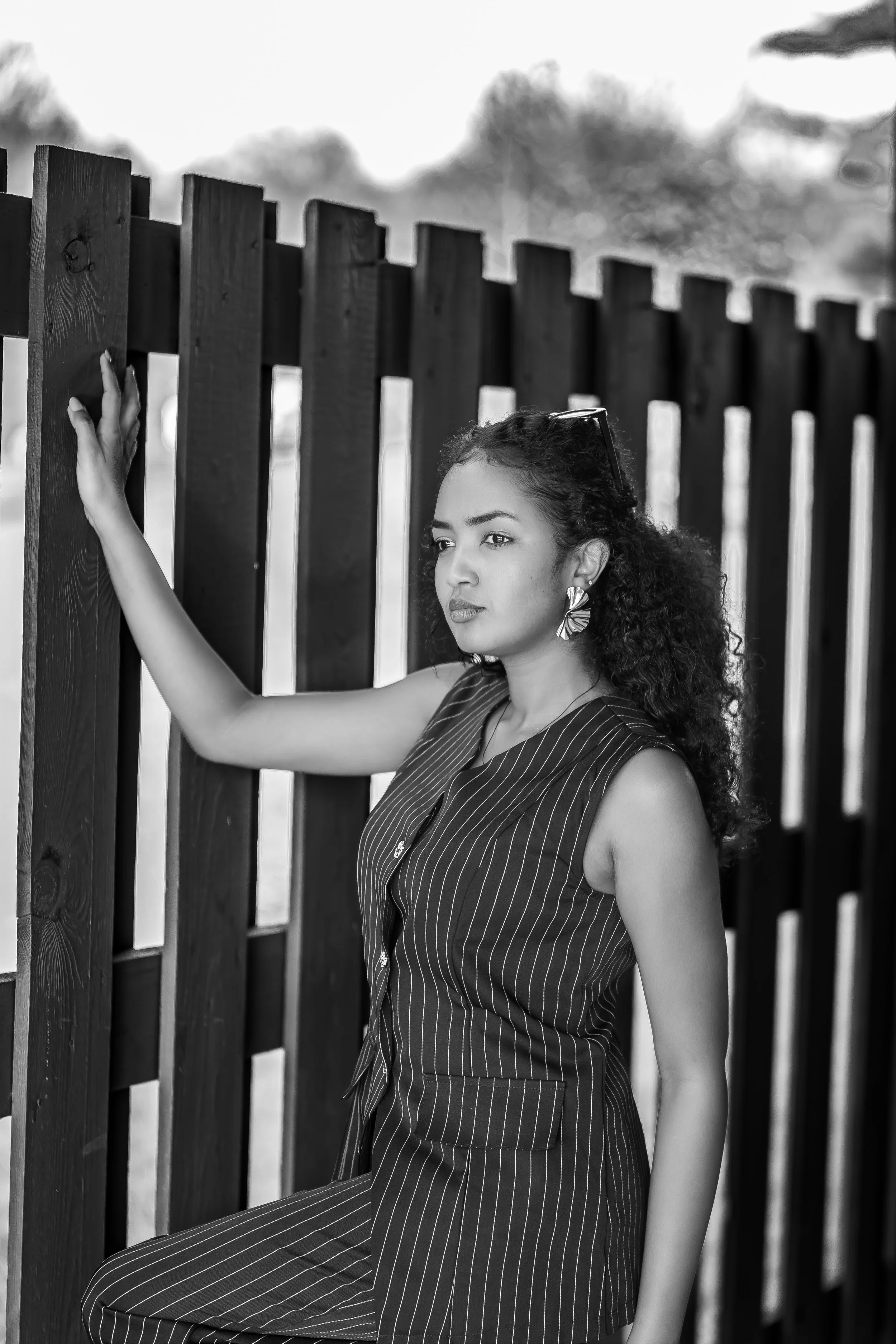 A young woman with curly hair and large earrings, wearing a pinstriped sleeveless jumpsuit, stands by a wooden fence, gazing thoughtfully into the distance.