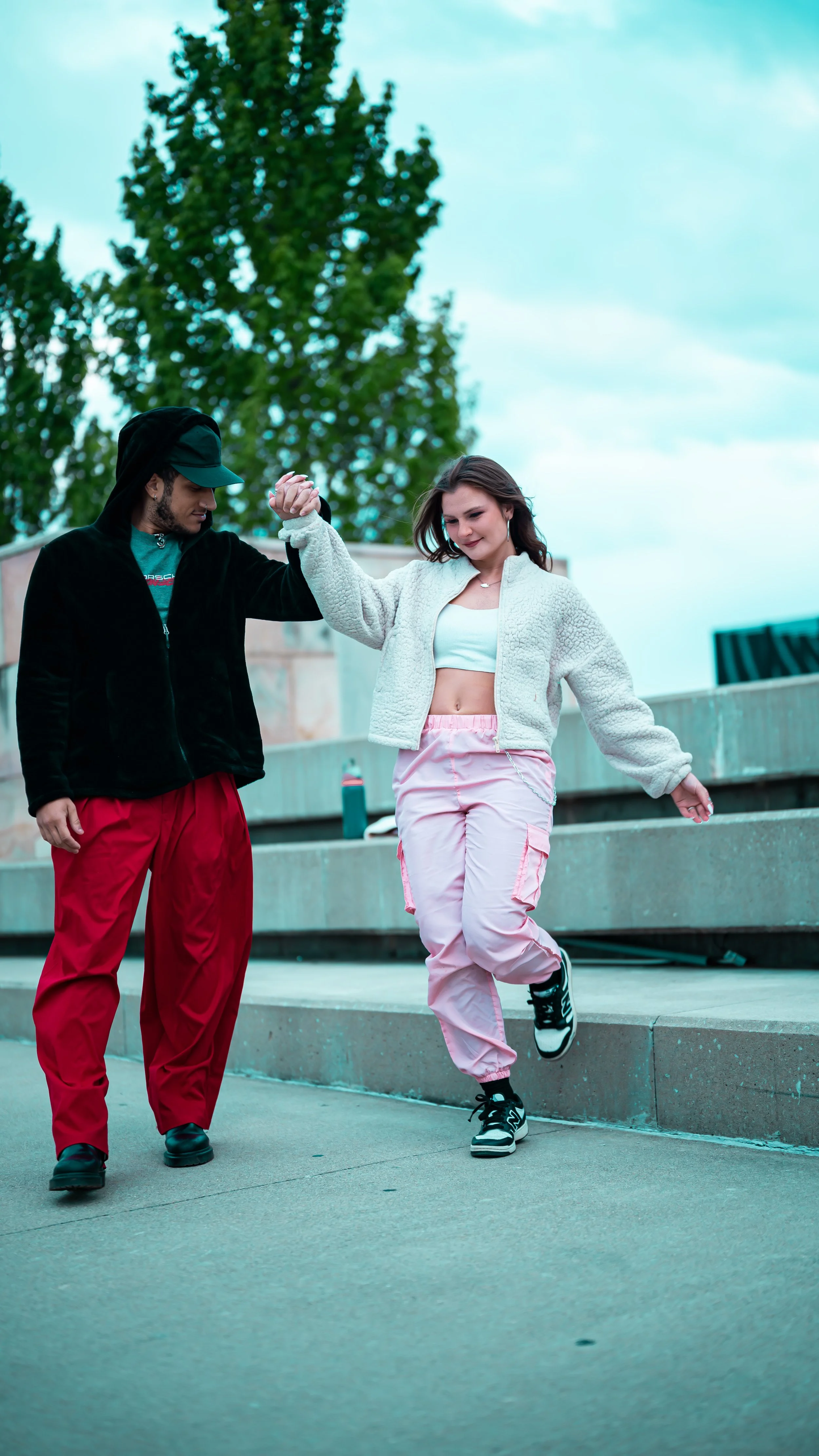 A man and woman dancing outdoors on a city sidewalk, holding hands, with trees and buildings in the background.