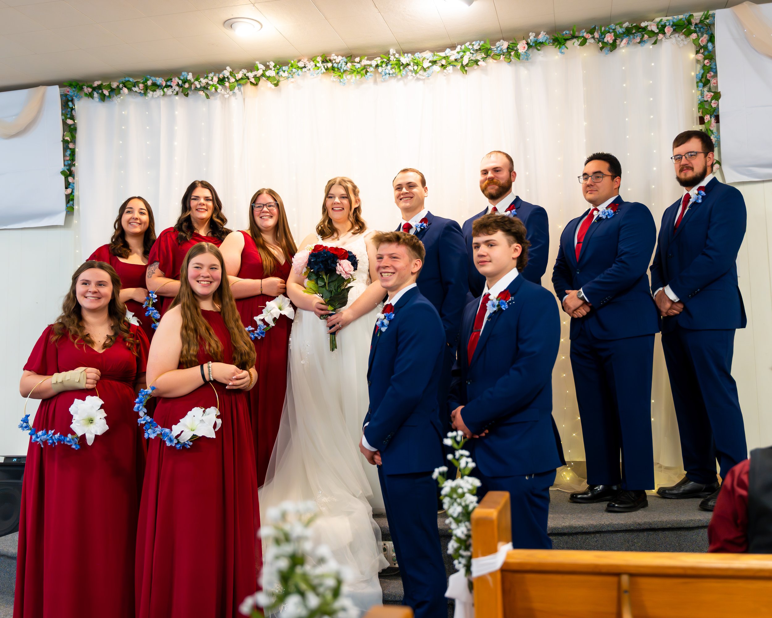 A wedding ceremony with the bride, groom, bridesmaids, and groomsmen standing on a decorated stage, smiling for the photo, with a white backdrop adorned with flowers and string lights.
