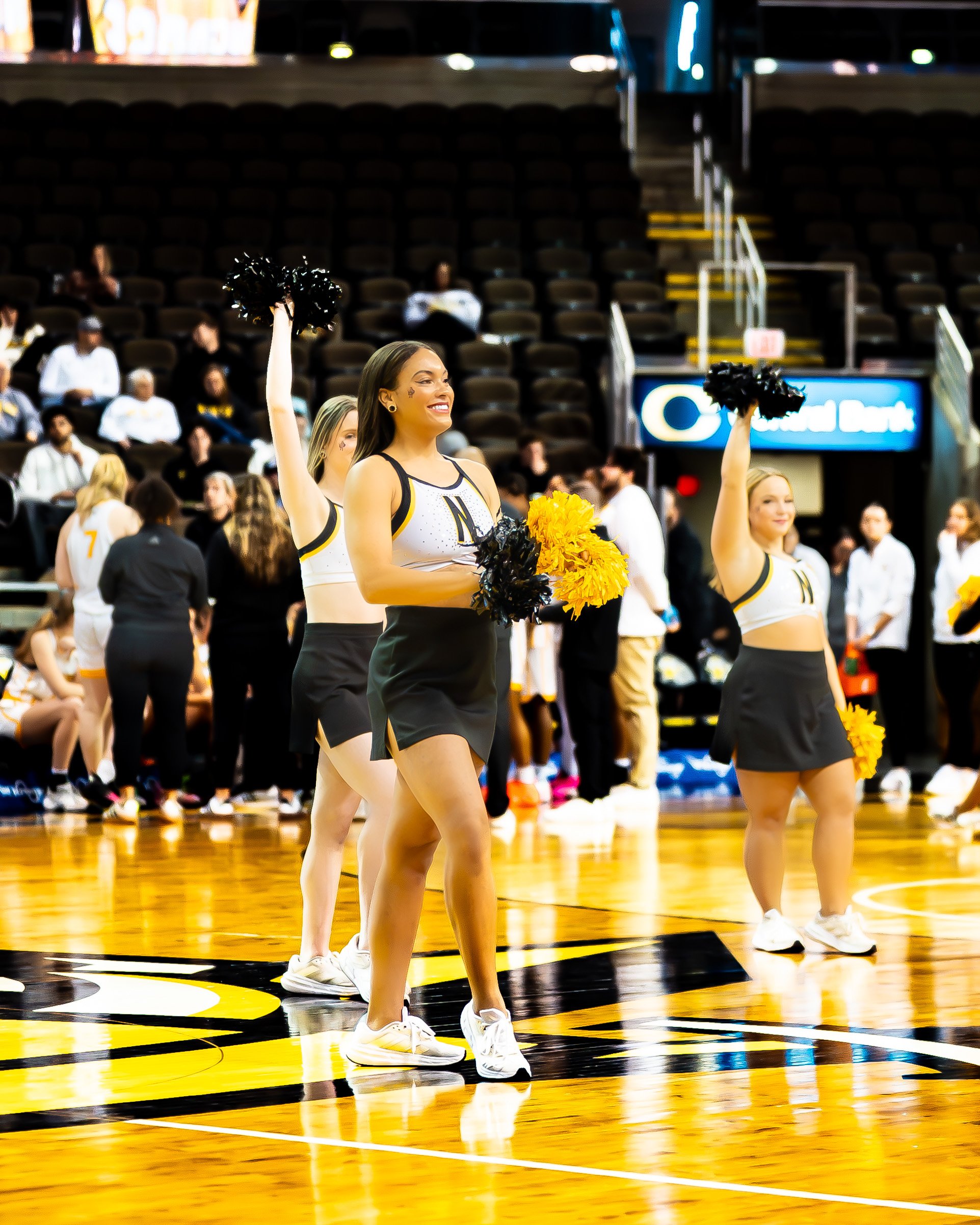 Cheerleaders performing on a basketball court, holding black and yellow pom-poms, with a mostly empty stadium in the background.
