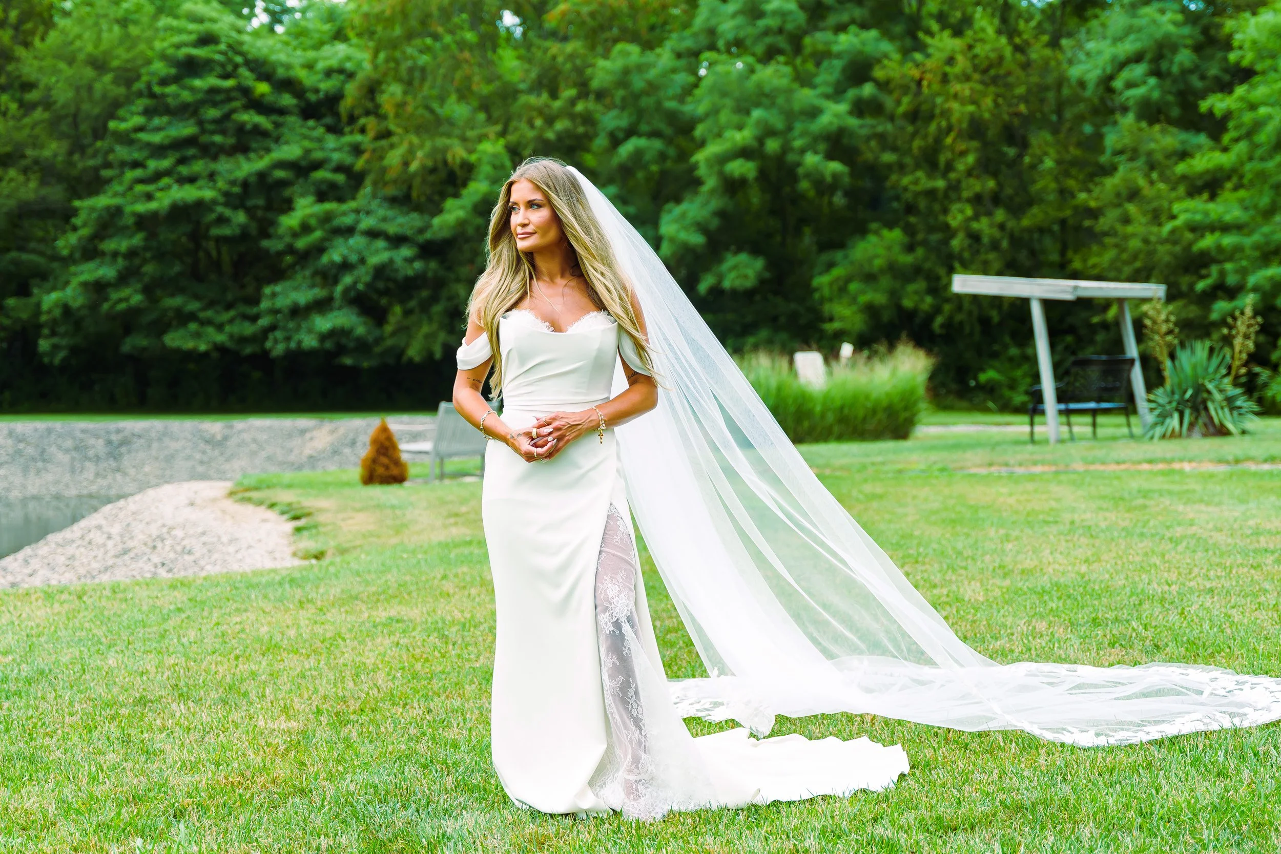 A woman in a white wedding dress with a long train and veil standing outdoors on a grassy area, with green trees in the background.