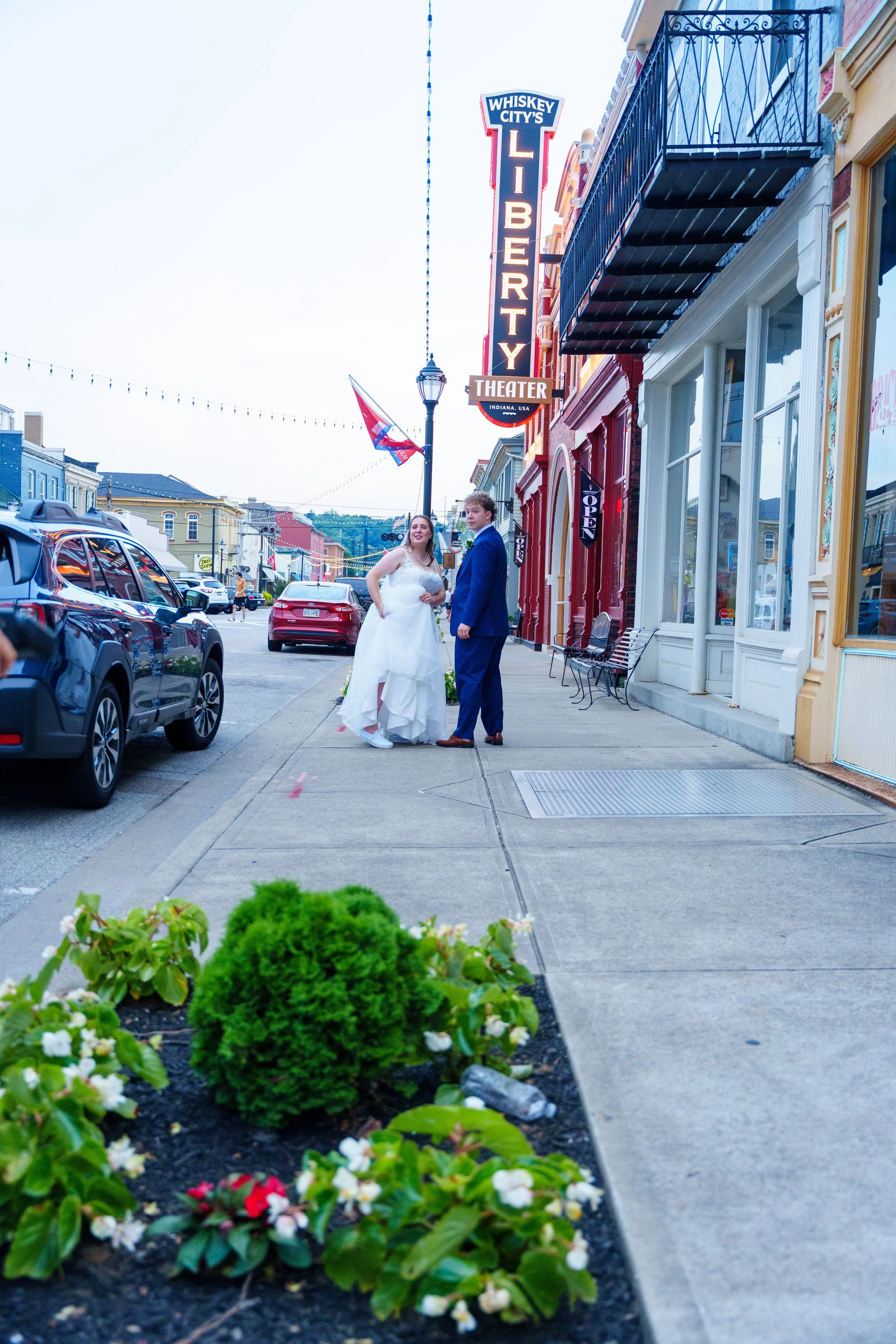 A bride and groom standing on a city sidewalk in front of a theater with neon signs, parked cars, and storefronts on both sides, with a flower bed in the foreground.
