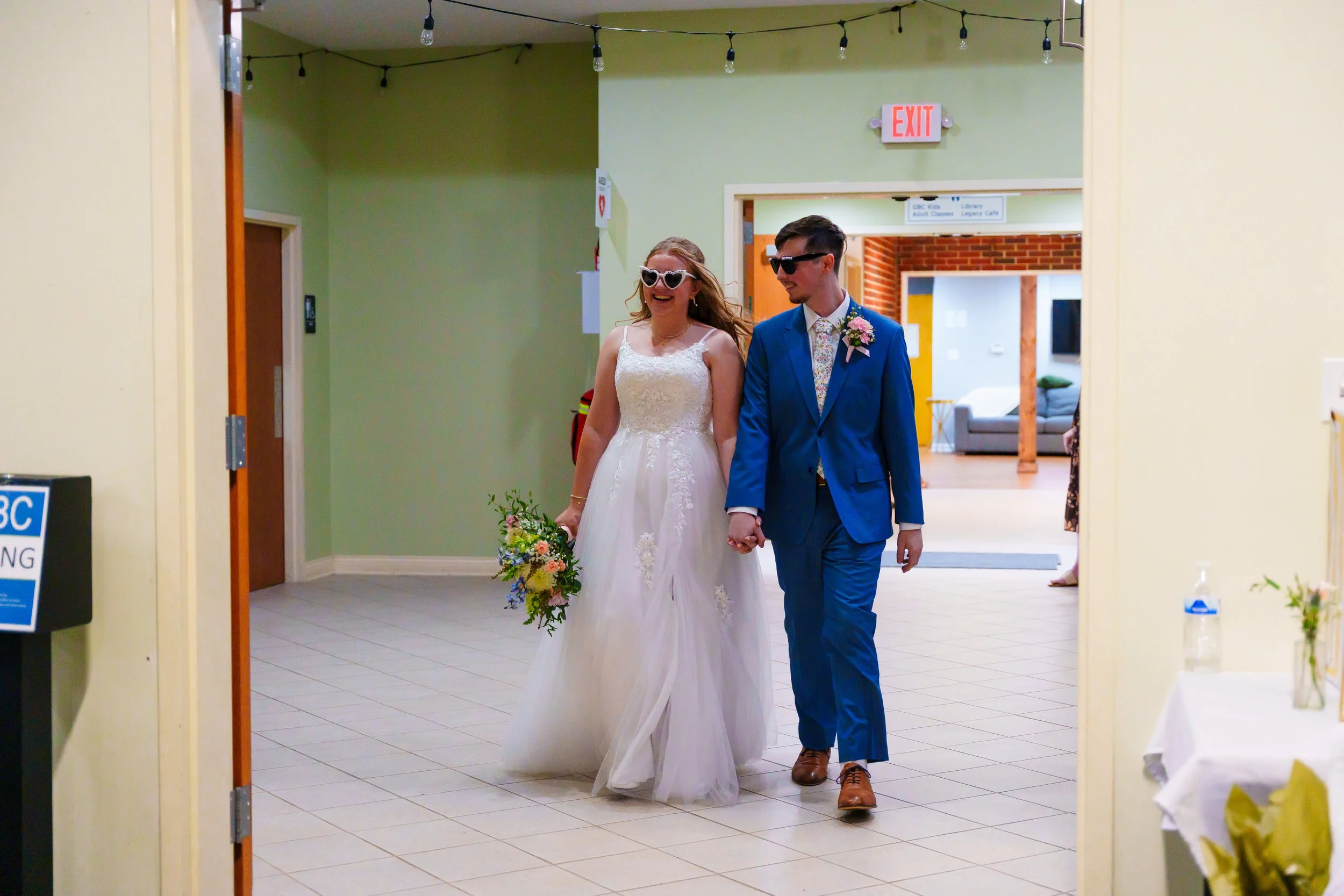 A bride and groom walking together in a brightly decorated indoor setting, holding hands. The bride is wearing a white lace wedding dress and sunglasses, holding a colorful bouquet. The groom is in a bright blue suit with sunglasses, smiling at the b