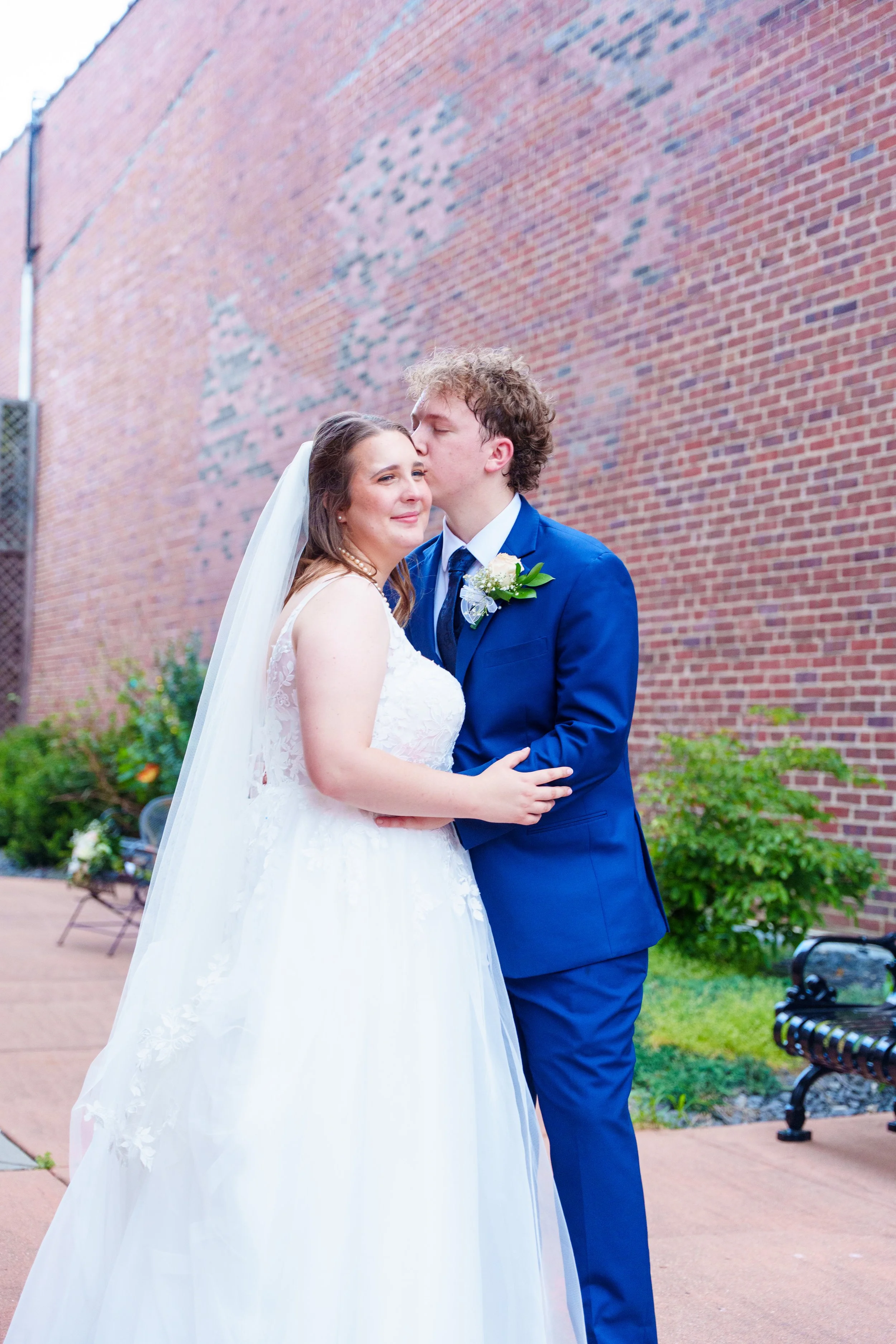 A bride and groom sharing a kiss outdoors near a brick wall. The bride is wearing a white wedding gown and veil, and the groom is in a blue suit with a white shirt and a boutonnière. The groom is kissing the bride on her forehead, and the bride is sm