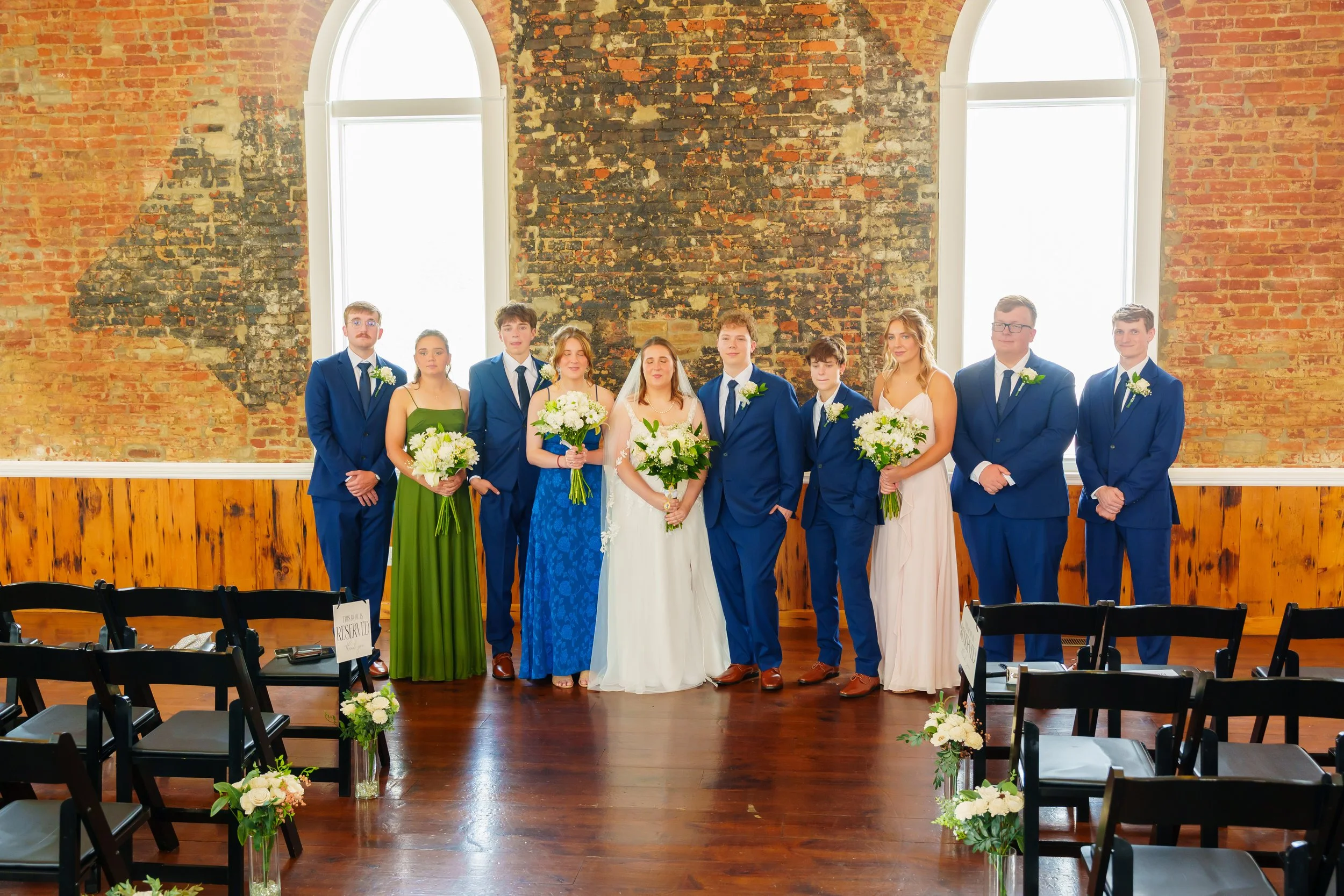A bride and groom with eight attendants, dressed in formal attire, standing in front of a brick wall with large arched windows during a wedding ceremony.