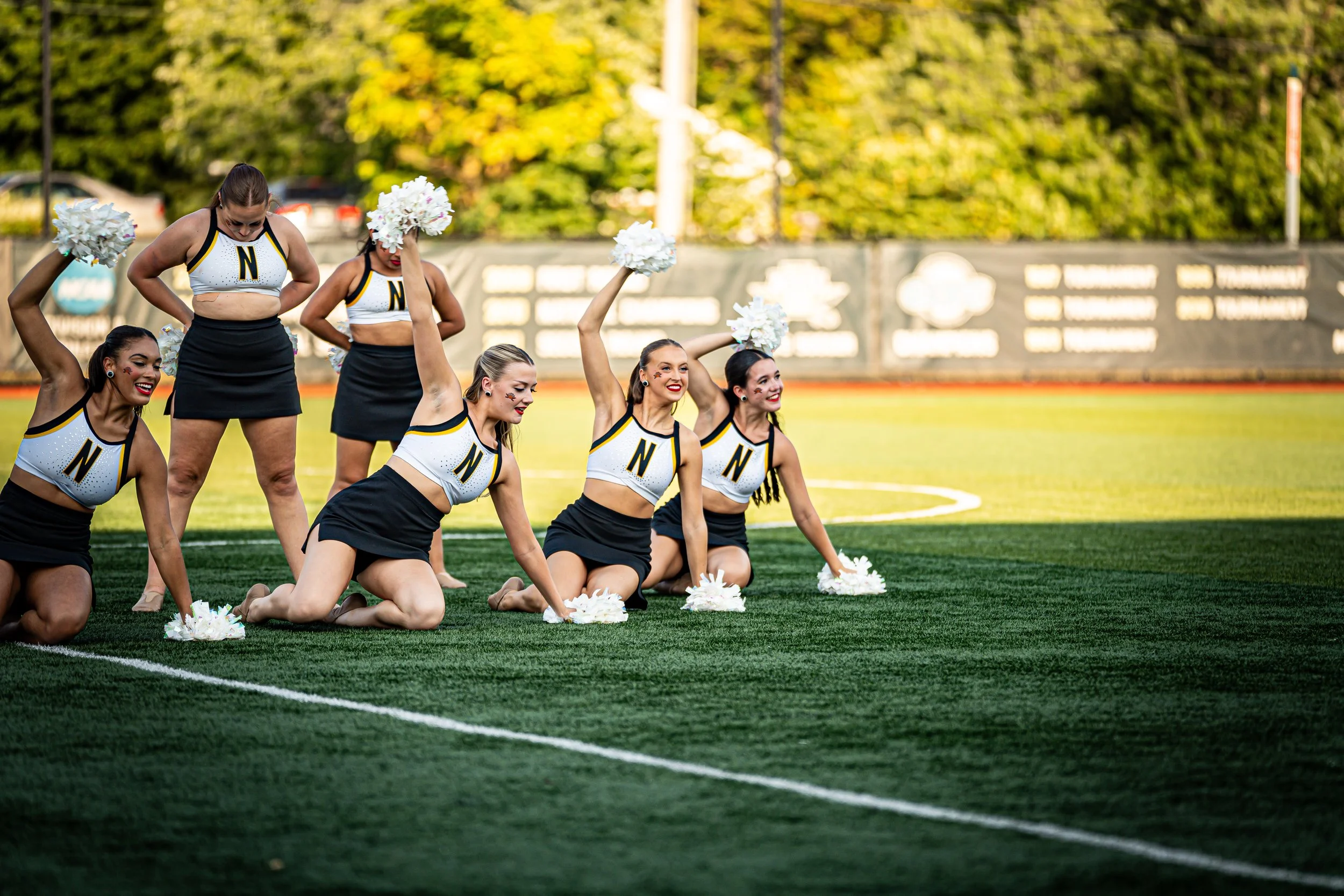 Group of cheerleaders in black and white uniforms with a letter 'N' posing on a football field, some kneeling and some standing, holding white pom-poms, with trees and banners in the background.
