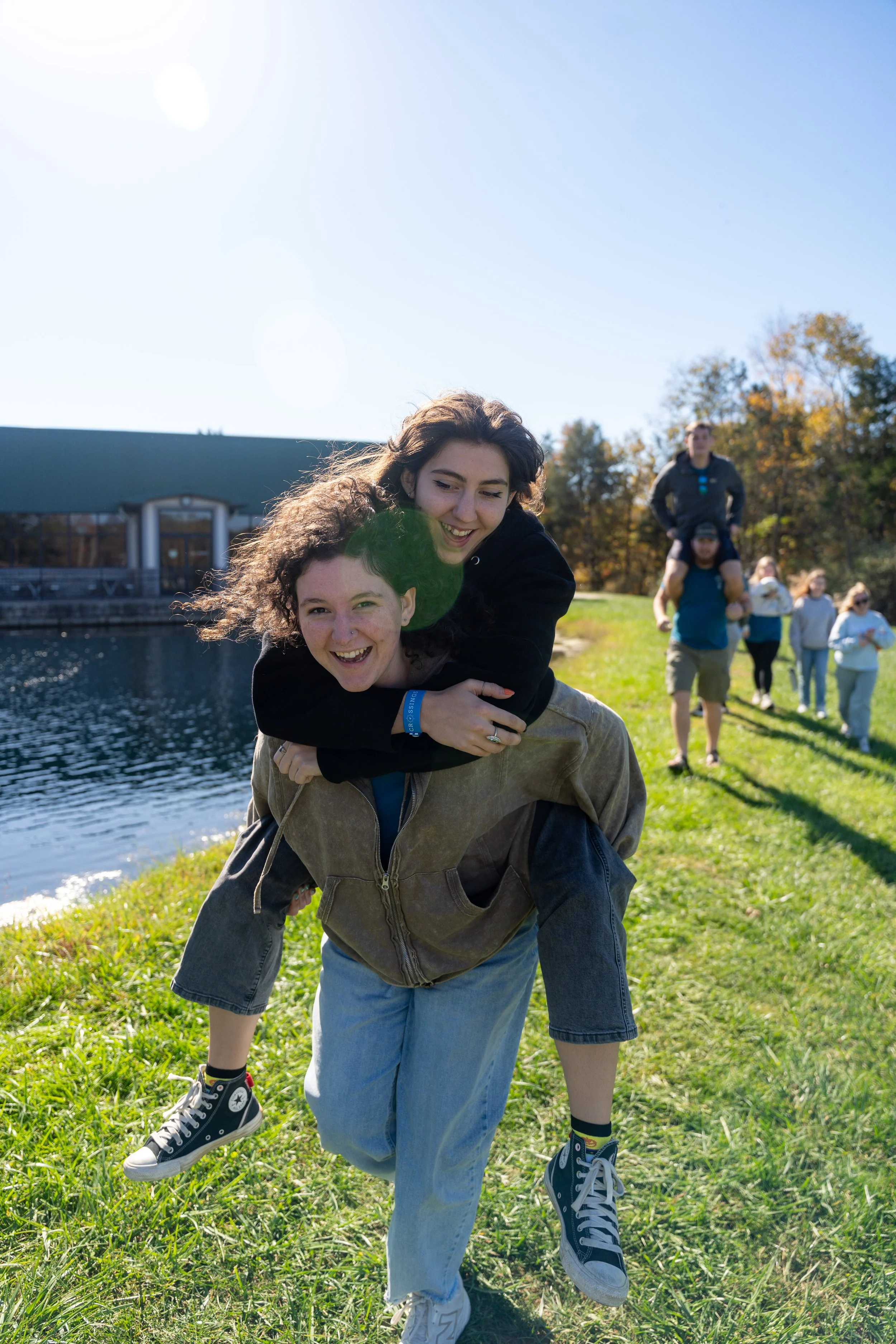 Two young women are outdoors, one giving the other a piggyback ride by the water. The woman in front is wearing a brown jacket, jeans, and sneakers, while the woman on her back has curly hair and is smiling. In the background, a group of people is wa