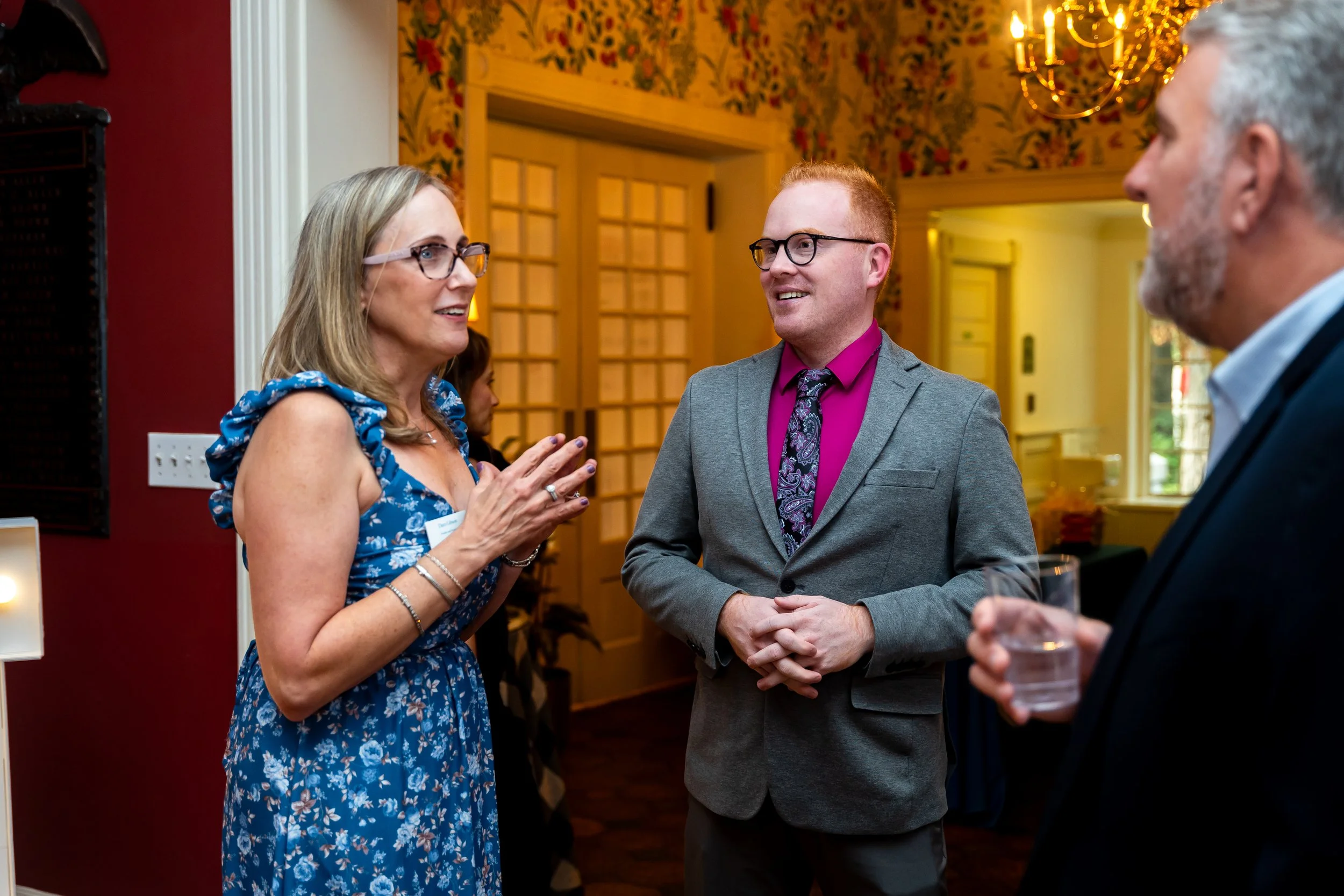 Three people engaged in a conversation at a social gathering indoors; a woman with glasses and a blue floral dress appears to be explaining something, a man with glasses and a gray suit listens attentively, another man with gray hair and a suit holds