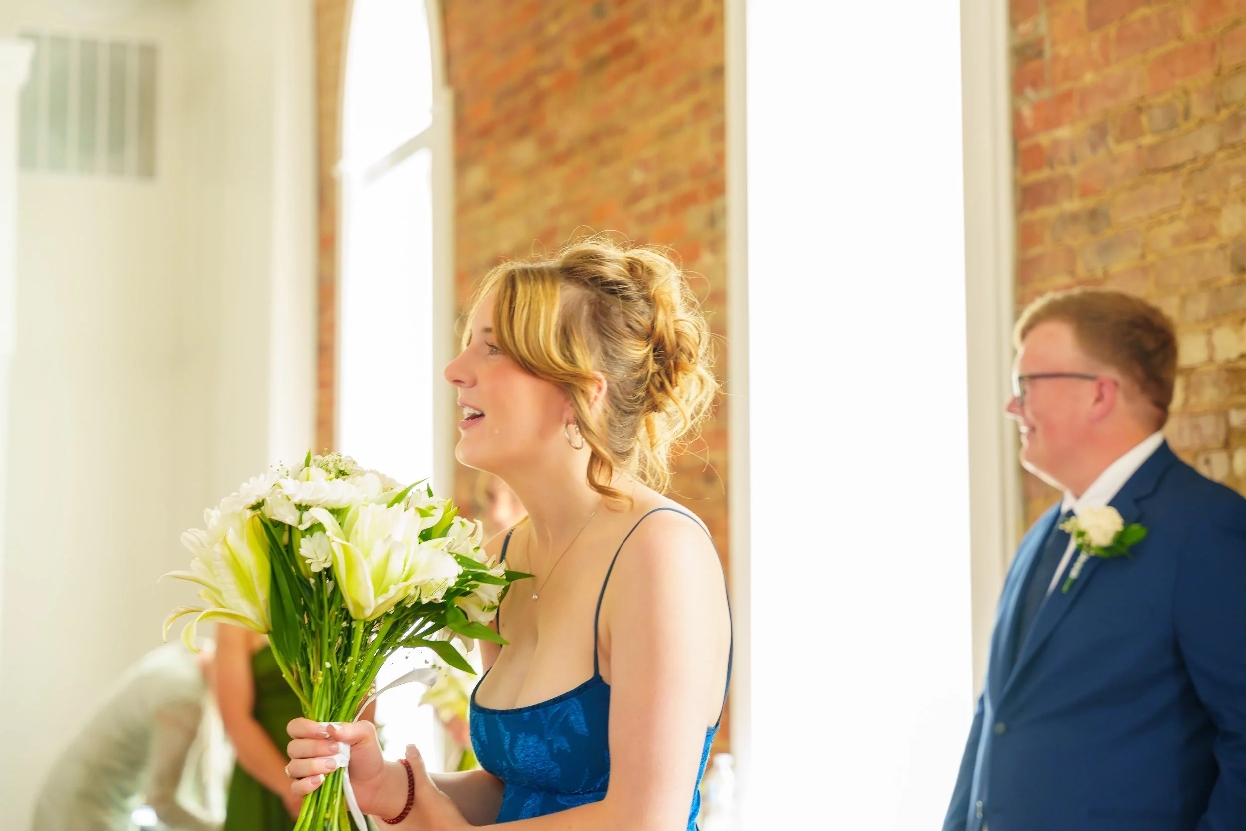 A woman in a blue dress holds a bouquet of white lilies with greenery, standing in a room with a brick wall and large windows, at a wedding or special event.