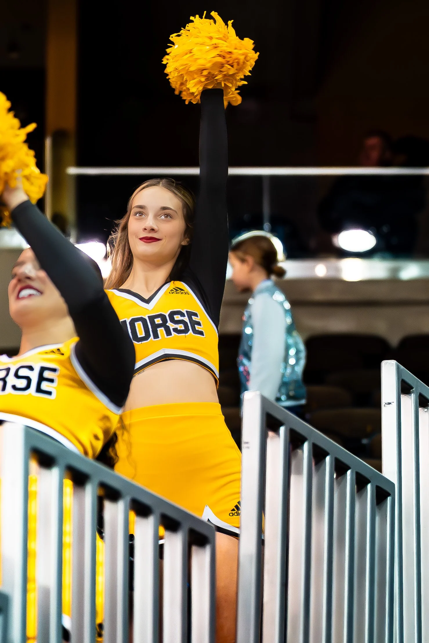Cheerleaders in yellow and black uniforms performing at a sports event, with one cheerleader raising her pom-pom.