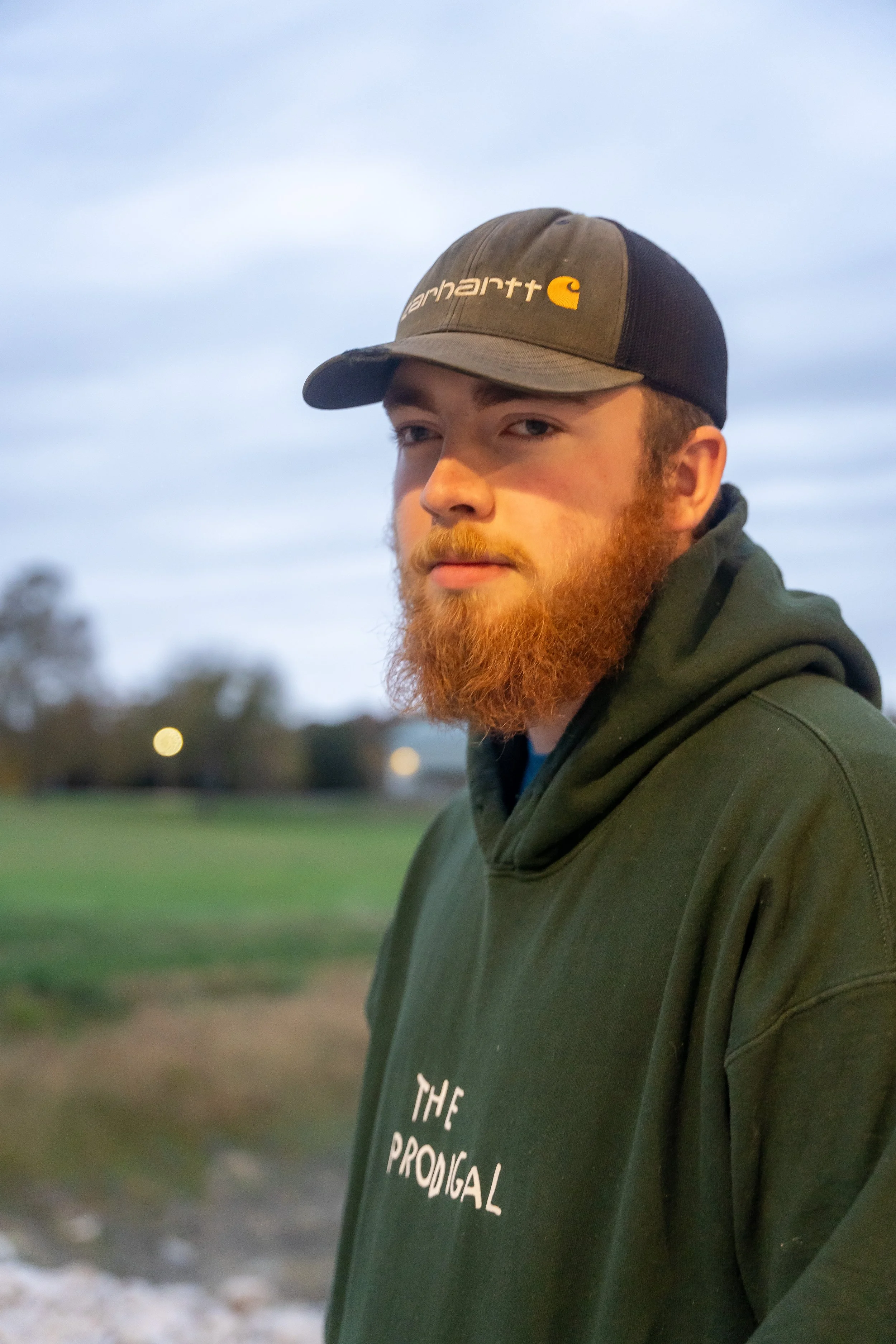 A young man with a red beard and mustache wearing a khaki and black Carhartt baseball cap, standing outdoors with trees and grass in the background during evening.