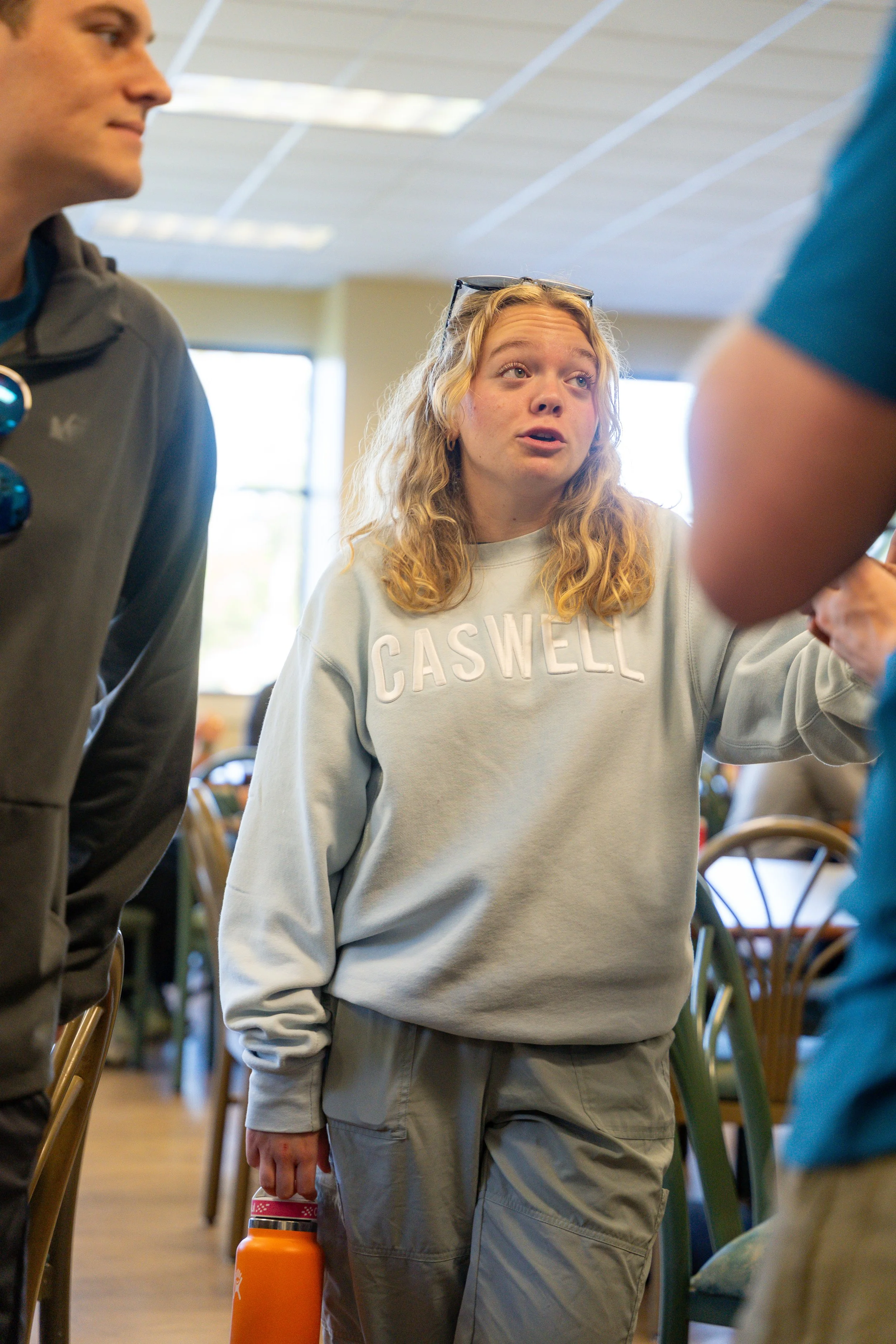 A young woman with blonde curly hair wearing a light gray sweatshirt with the word 'CASWELL' and gray pants, standing indoors surrounded by other people, holding an orange water bottle in her right hand.