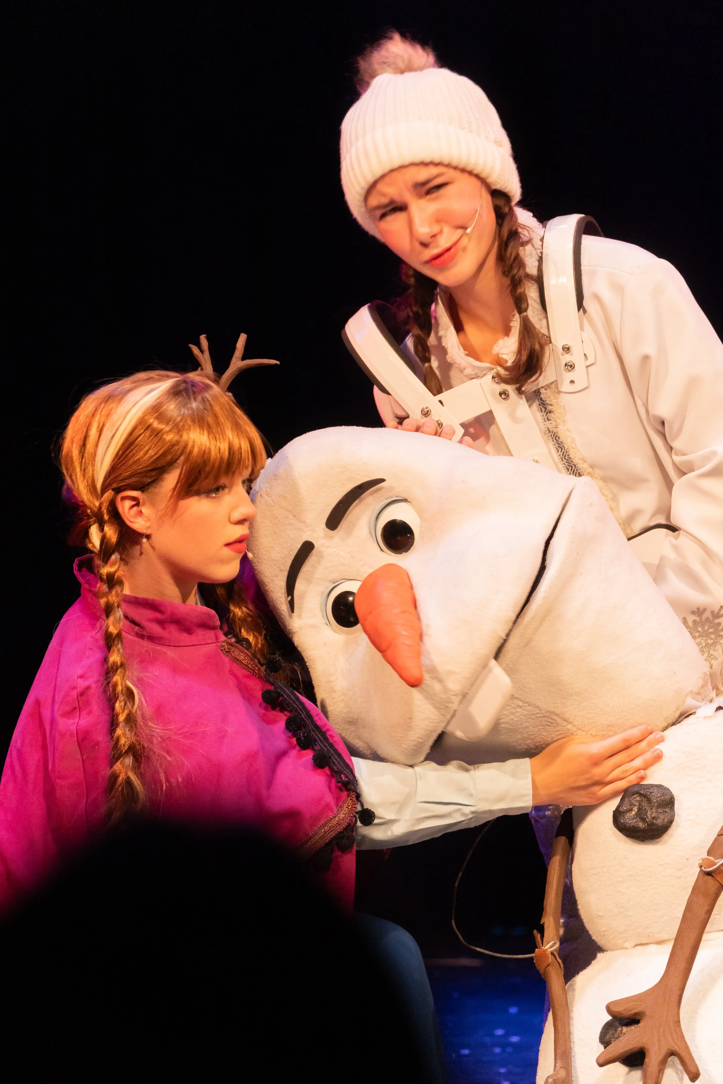 The image shows two women and a large snowman figure on stage. The woman on the left has long blonde hair in braids, wearing a purple jacket, and is looking at the snowman. The woman on the right has dark hair in braids, wearing a white coat and knit