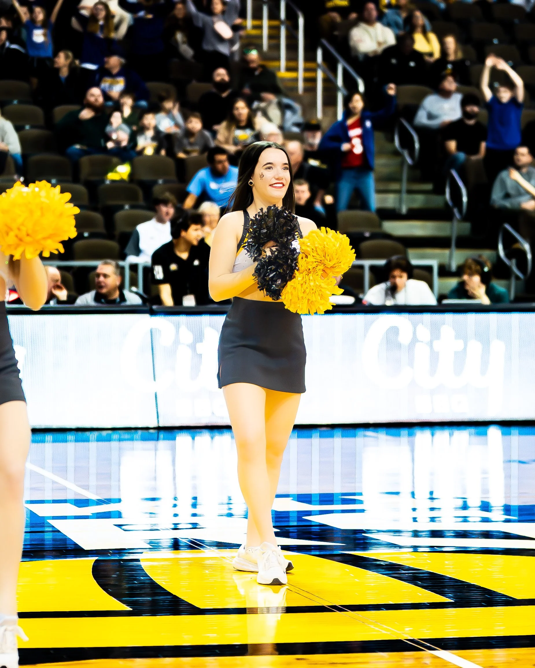 A cheerleader holding yellow and black pom-poms stands on a basketball court, smiling, with spectators in the background.