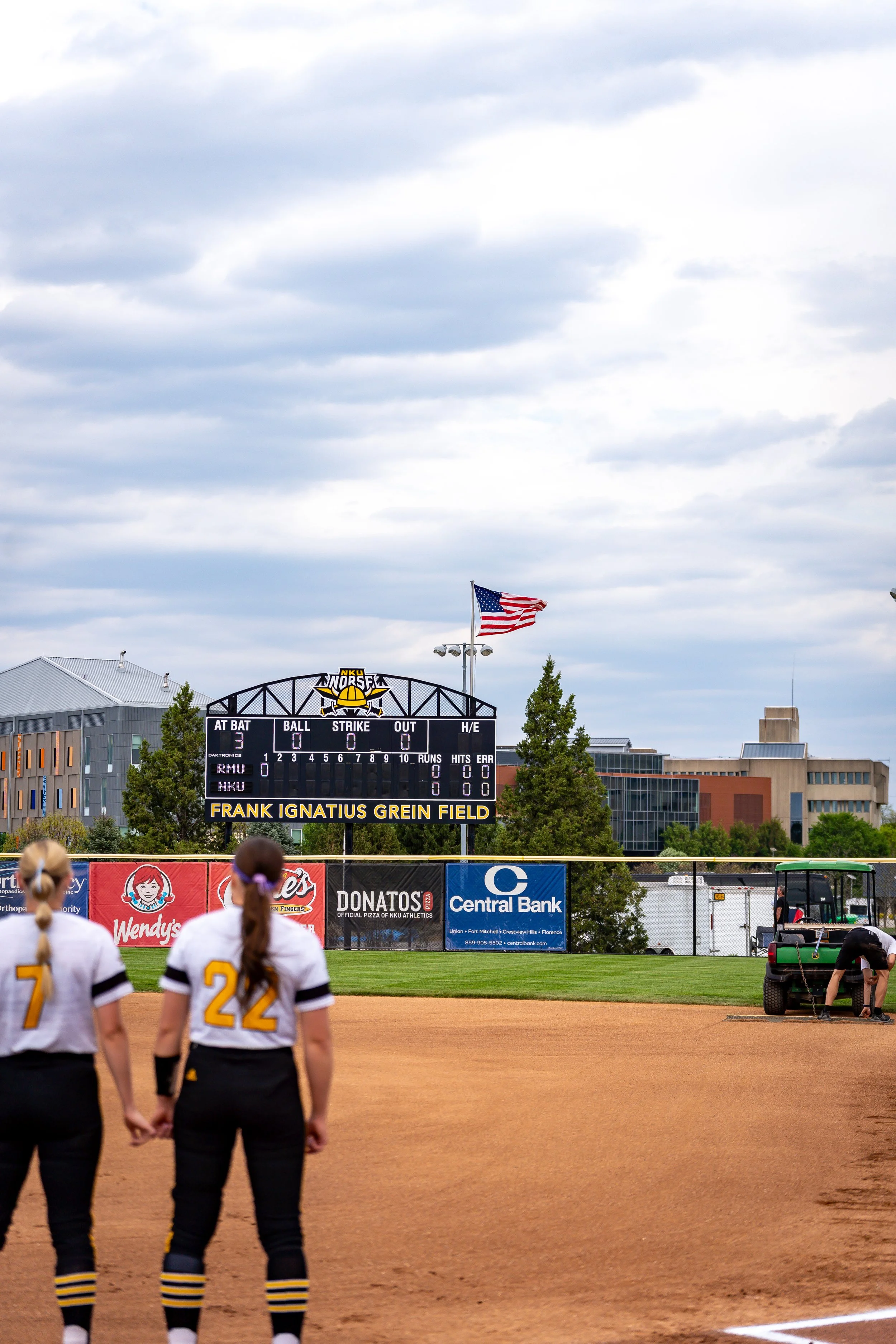 Sports field with players in black and yellow uniforms, football goal, and scoreboard displaying no scores, with American flag flying in the background under a cloudy sky.