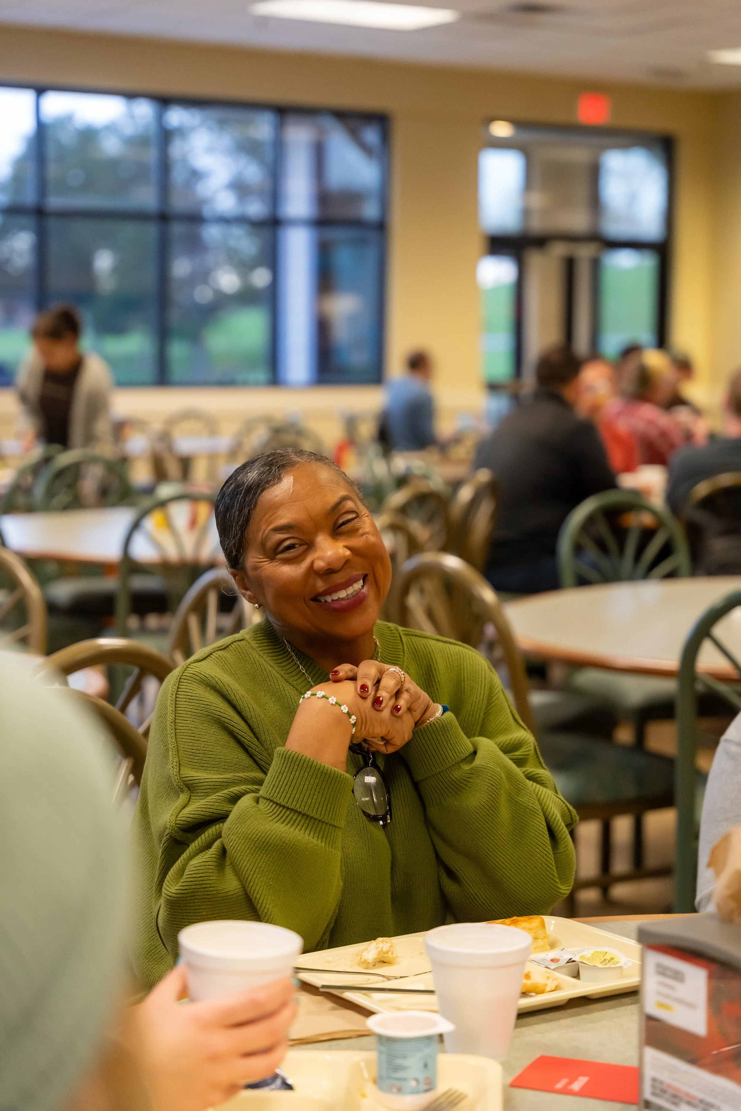 A woman smiling and sitting at a table in a cafeteria with other people in the background. She is wearing a green sweater and has her hands clasped together near her face.