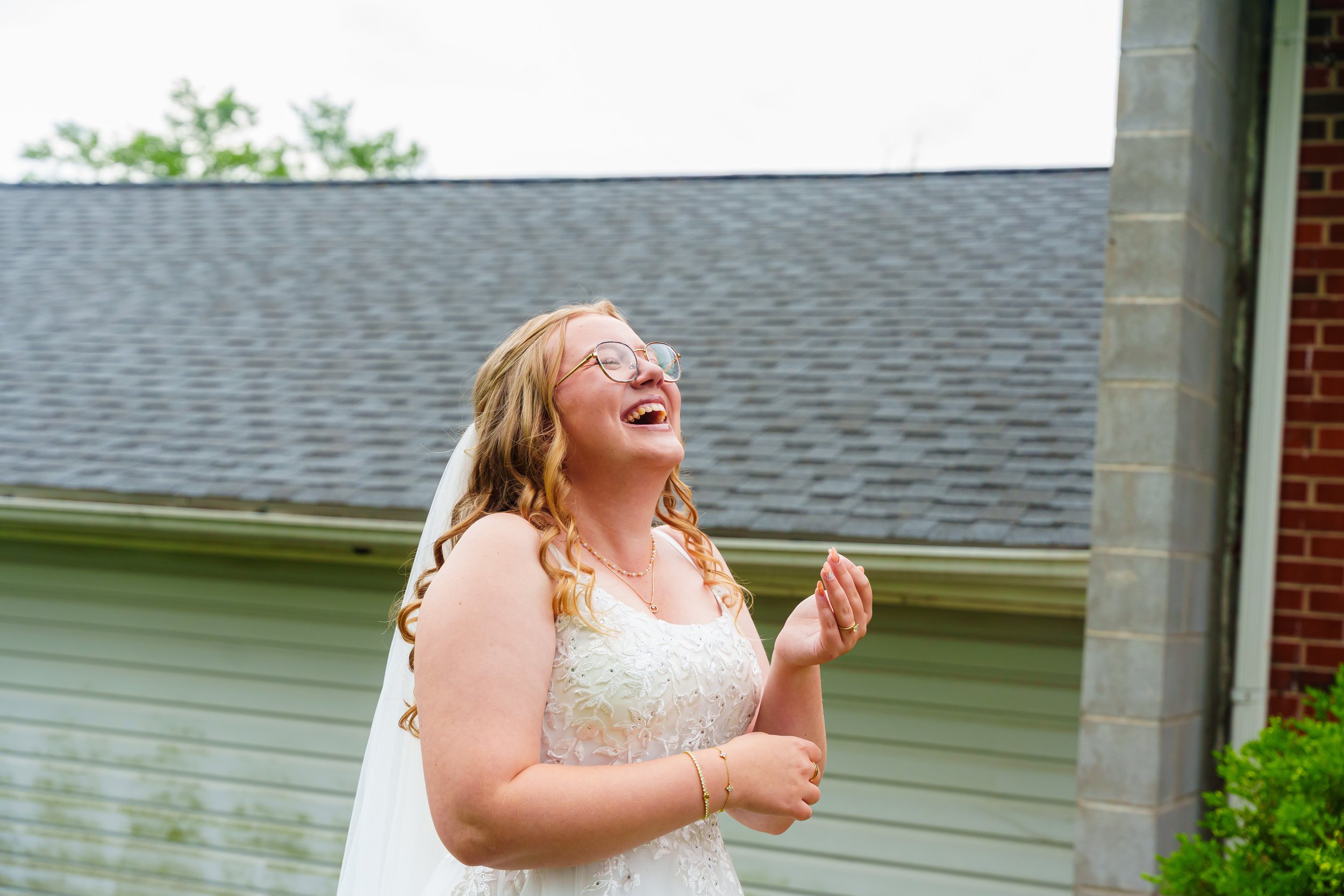 A woman with long, curly red hair, wearing glasses, a pearl necklace, and a white wedding dress, smiling and looking upward outdoors near a house with a gray shingled roof and brick chimney.