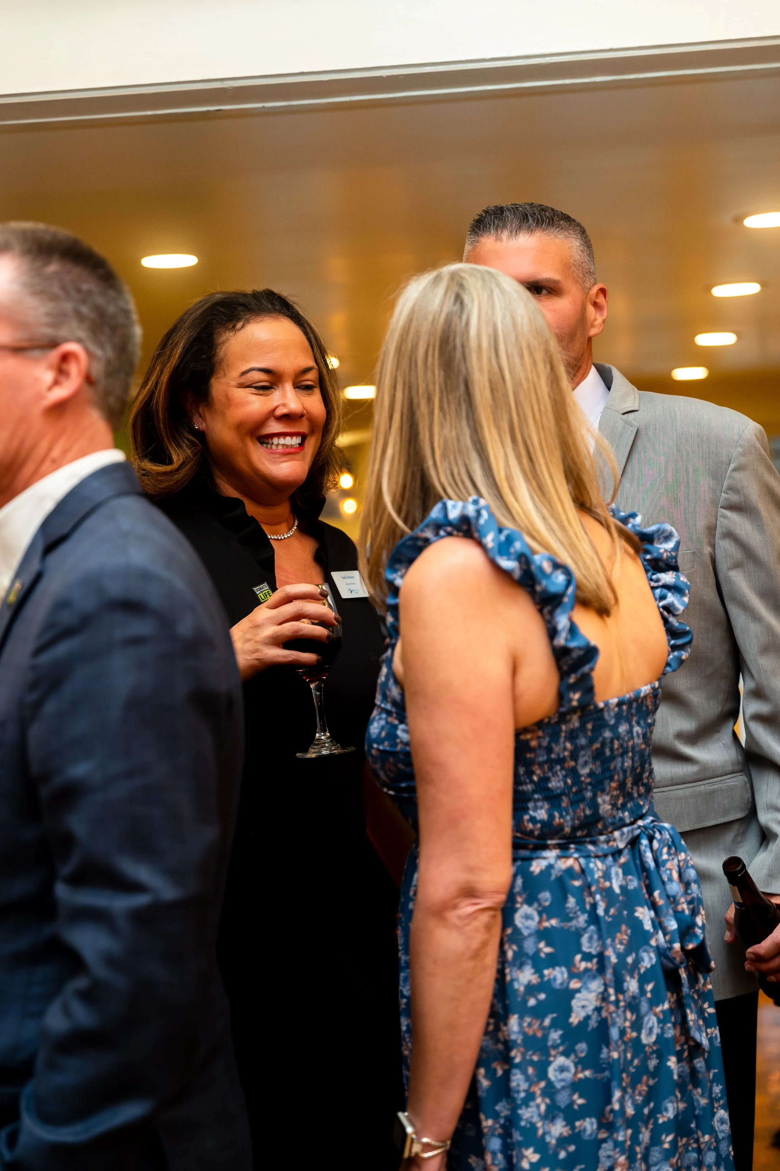 A group of people dressed in formal attire socializing at an indoor event. A woman in a black dress is smiling and holding a glass of wine, while others are engaged in conversation.