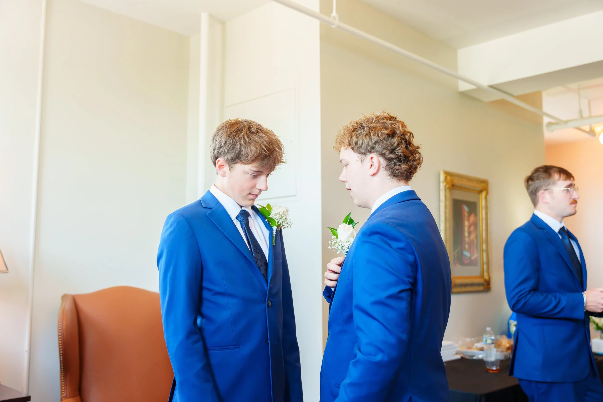 Two young men wearing blue suits and white shirts, with white boutonnieres, are standing close together in a room with cream-colored walls. They appear to be exchanging something or having a private moment. In the background, a third man in a blue su
